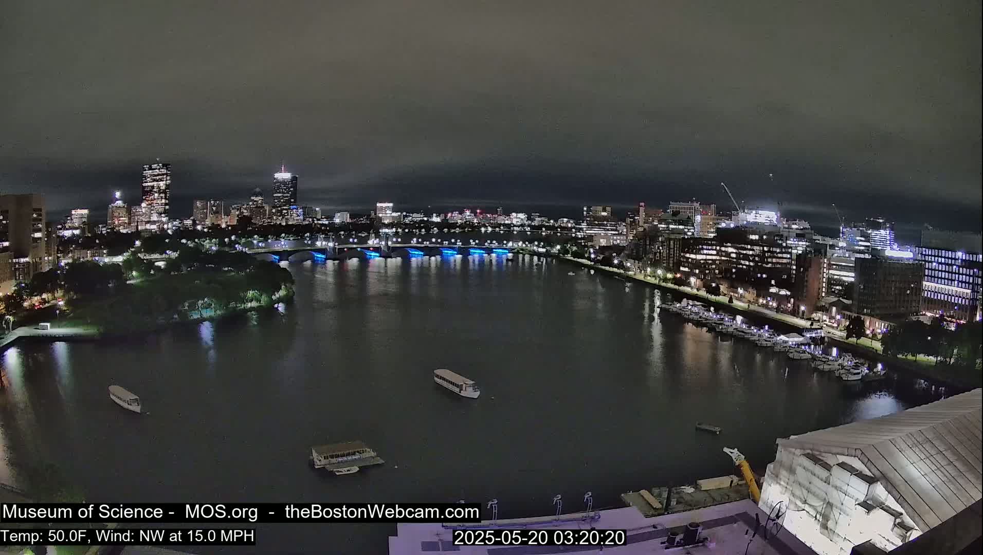 A nighttime view of a city skyline across a river, with boats docked along the shore and a bridge lit with blue lights, under a dark, cloudy sky with a 15 mph northwest wind and a temperature of 50°F.