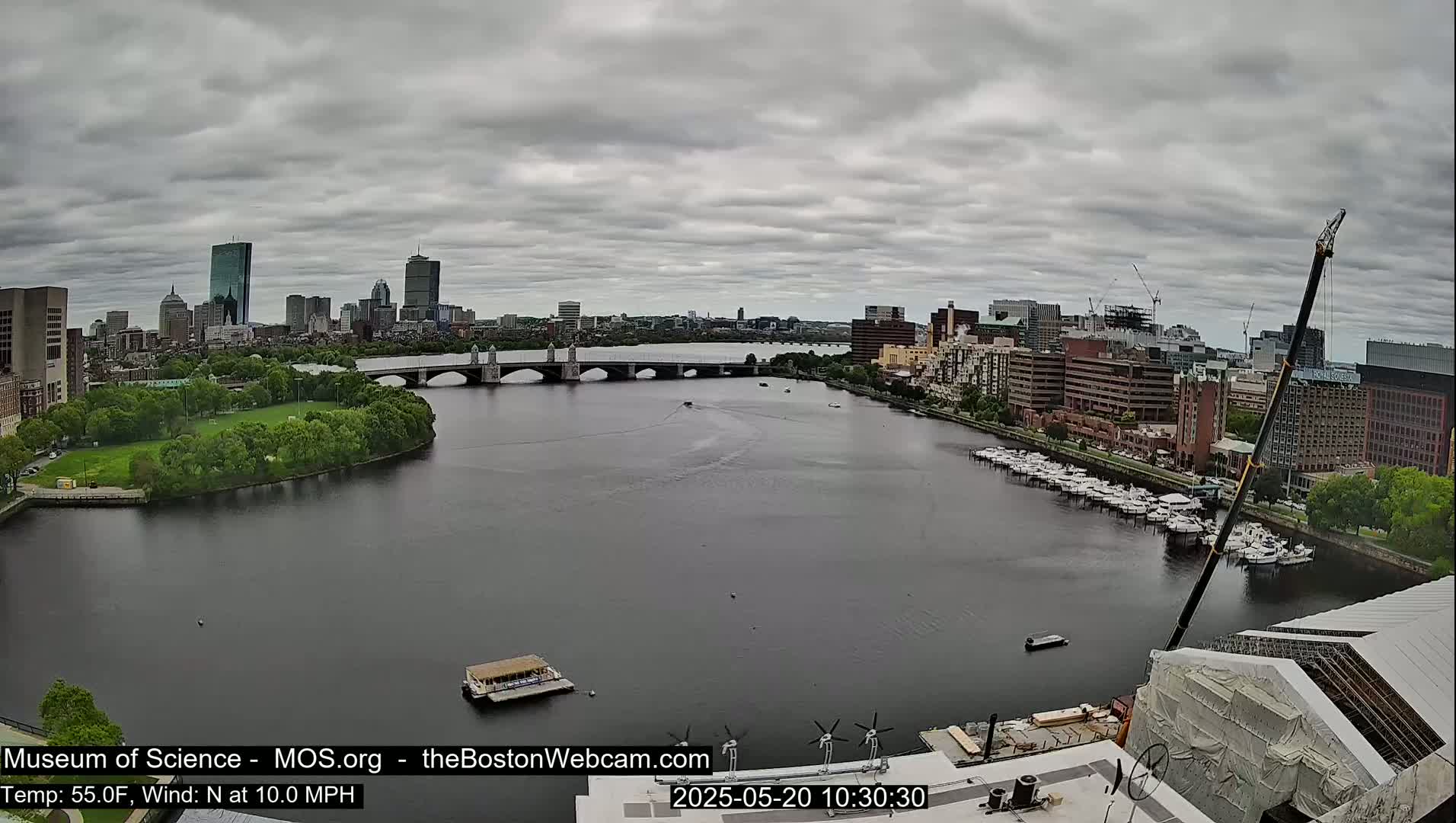 A city skyline is visible across a river, under a cloudy sky, with boats docked along the riverbank and a construction crane in the foreground.