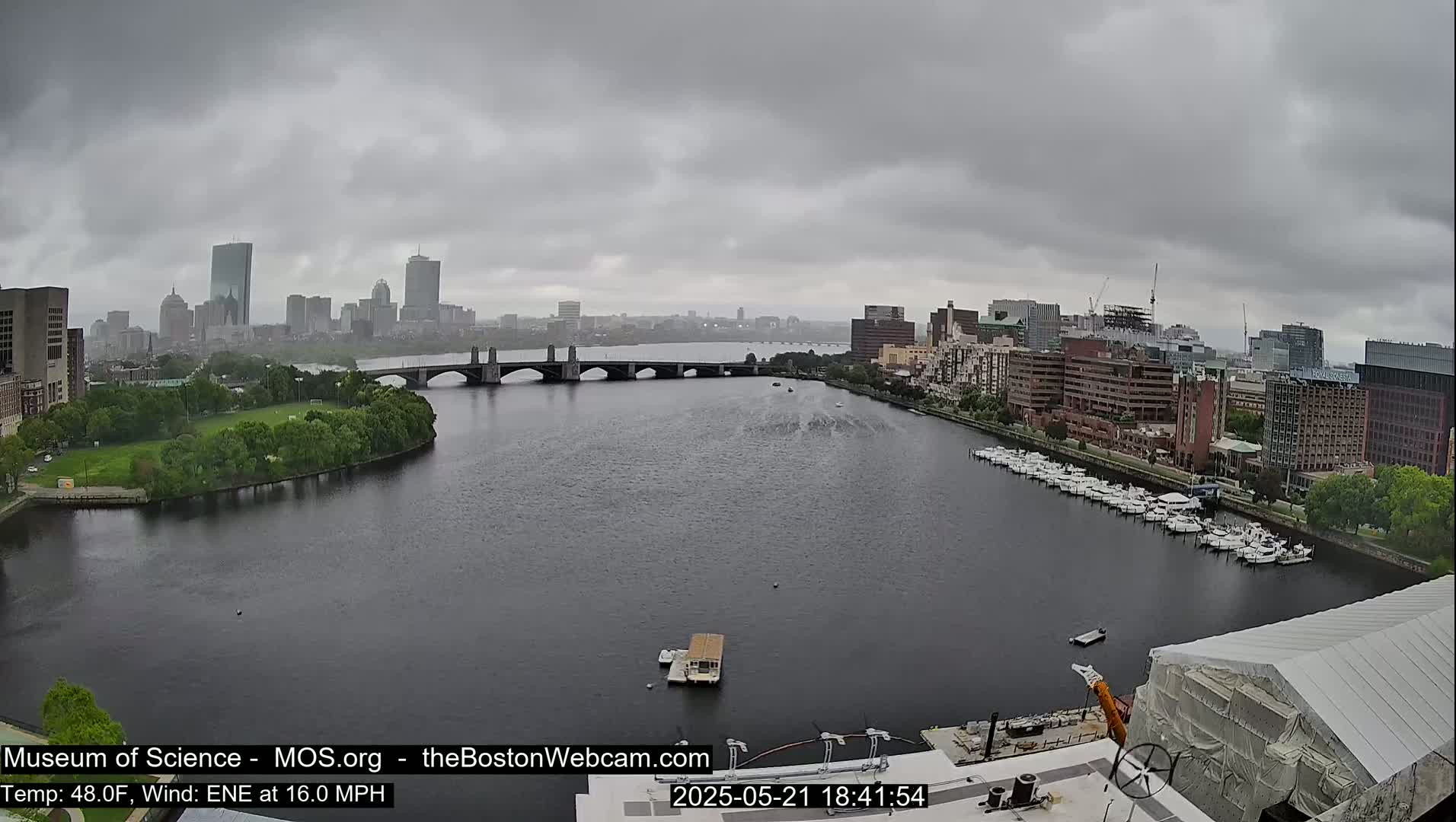 A city skyline under a gray, overcast sky is viewed from across a river, with a bridge and several docked boats visible.