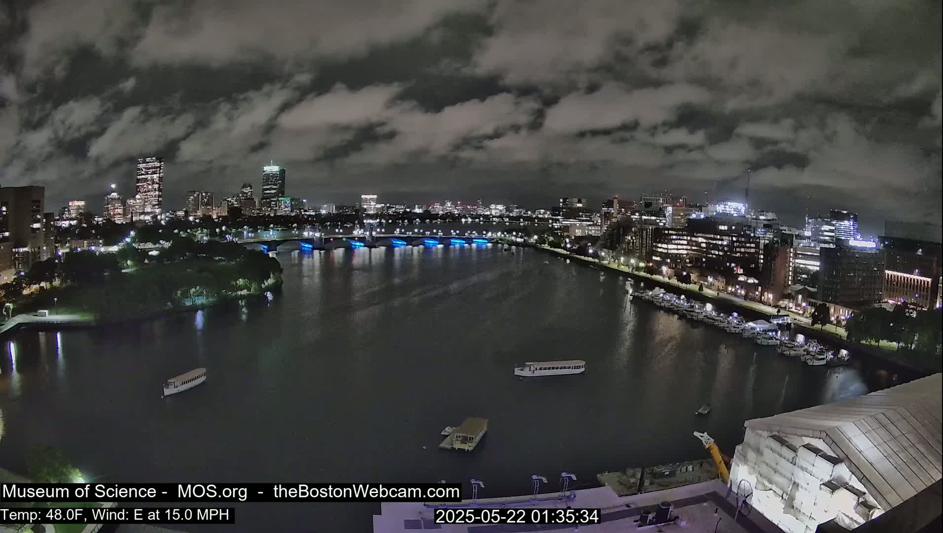 A nighttime cityscape is seen across a dark river, under a cloudy sky, with boats moored along the waterfront.