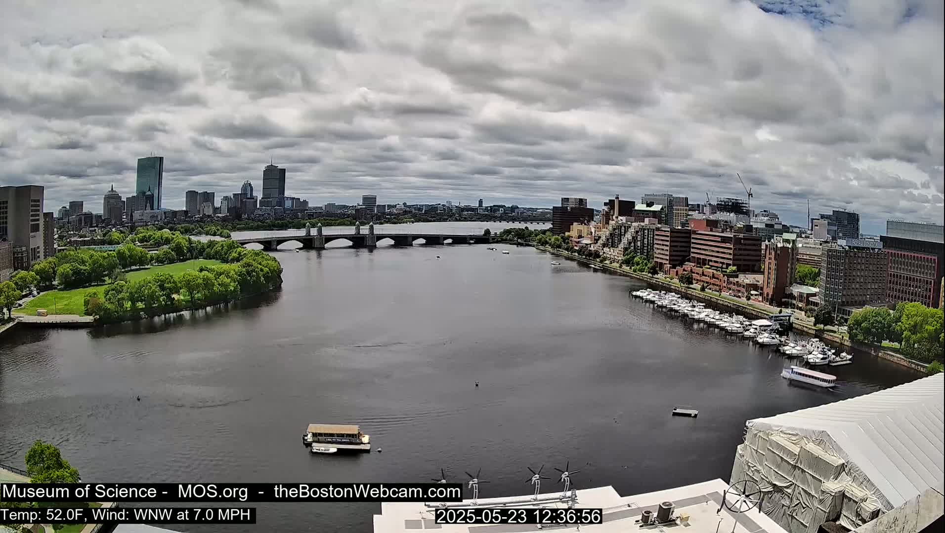 A wide shot of a river in a city, under a mostly cloudy sky, shows a bridge, boats, and a cityscape in the background.