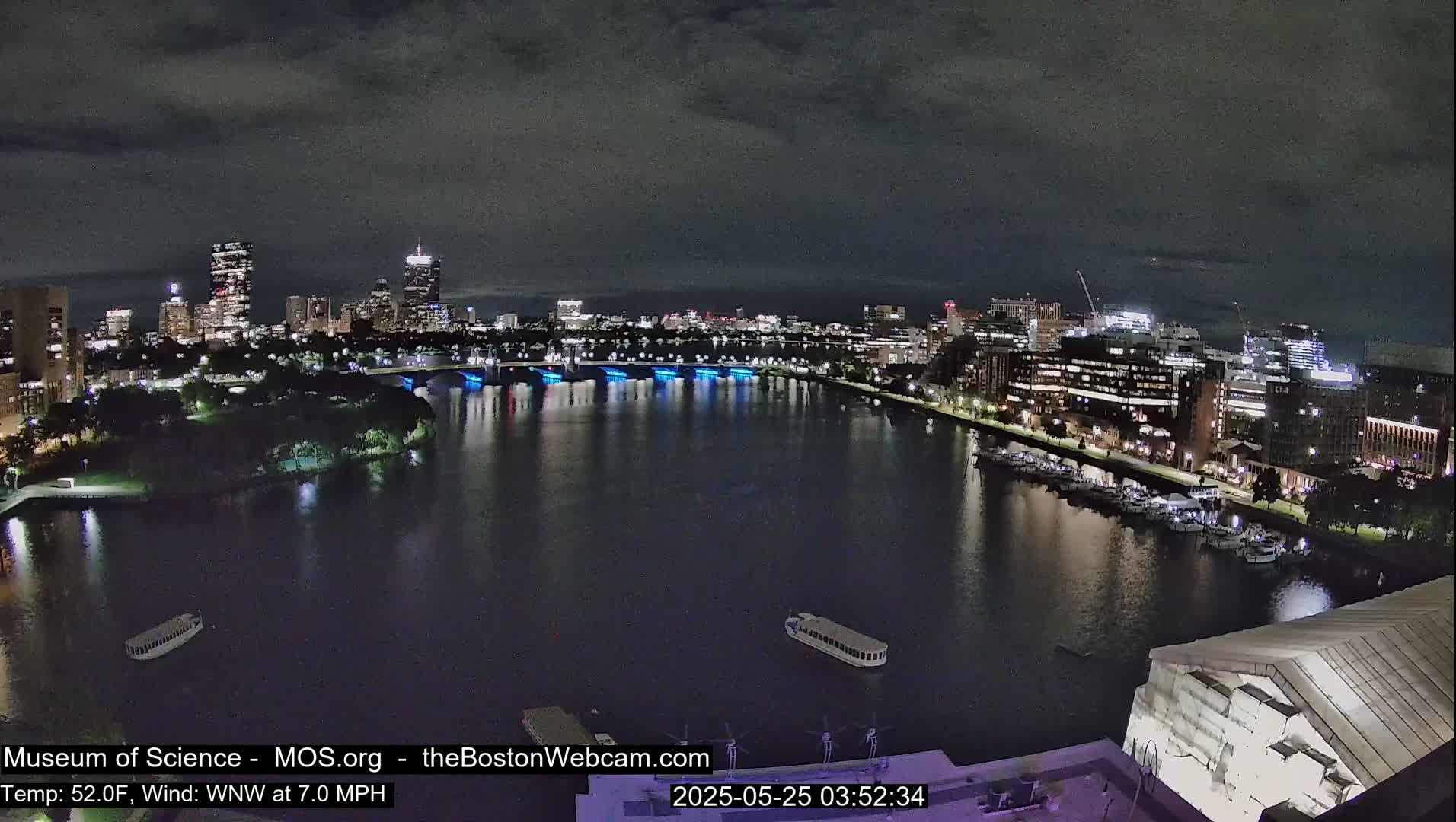 A nighttime view of a city skyline across a dark river, with boats moored along the shore under a cloudy sky.