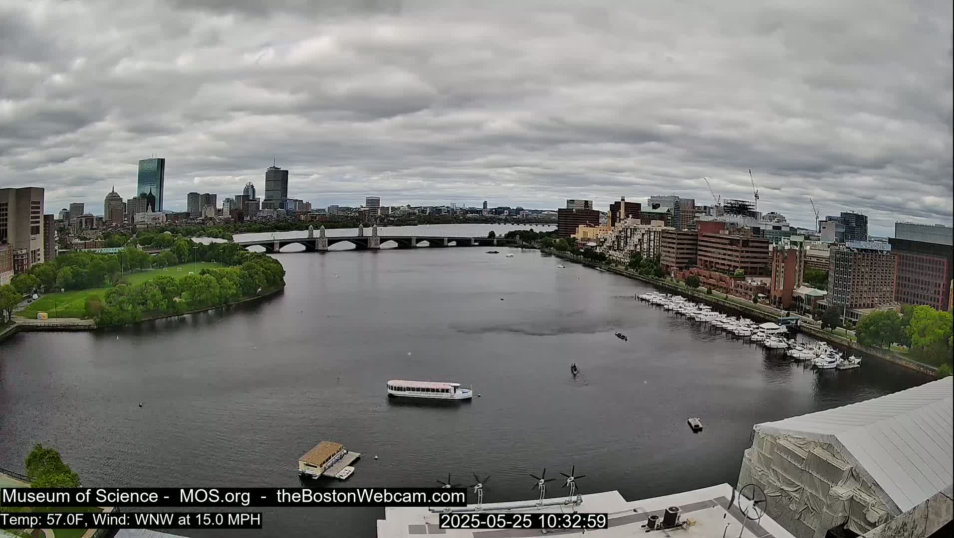 A wide shot of a city skyline across a river under a cloudy sky, showing a bridge, boats, and docked yachts.