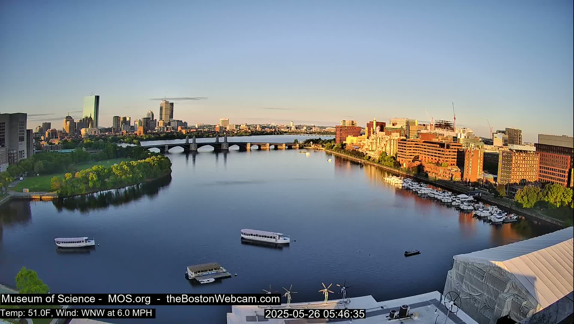 A city skyline is visible across a calm river under a clear, early morning sky, with several boats on the water and a bridge connecting the two sides.