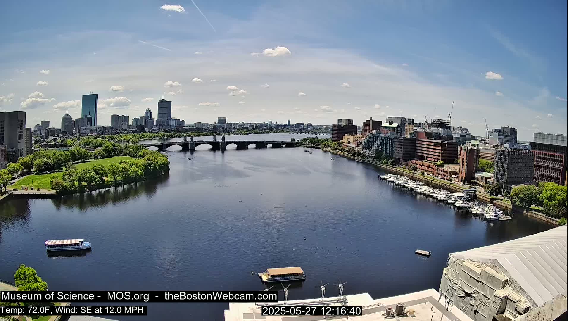 A wide shot of a city skyline across a calm river under a mostly sunny sky, with boats on the water and a bridge connecting the two sides.