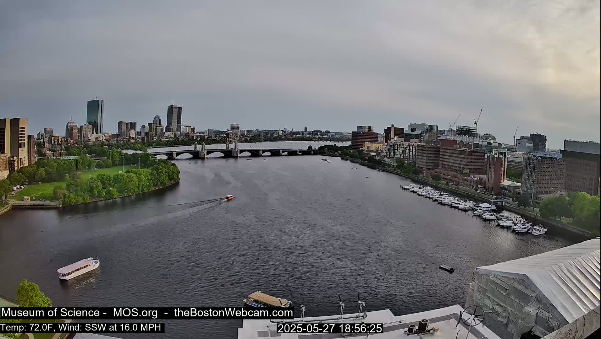 A wide shot of a city skyline across a river, under a mostly cloudy sky, shows a bridge, boats, and docked boats.