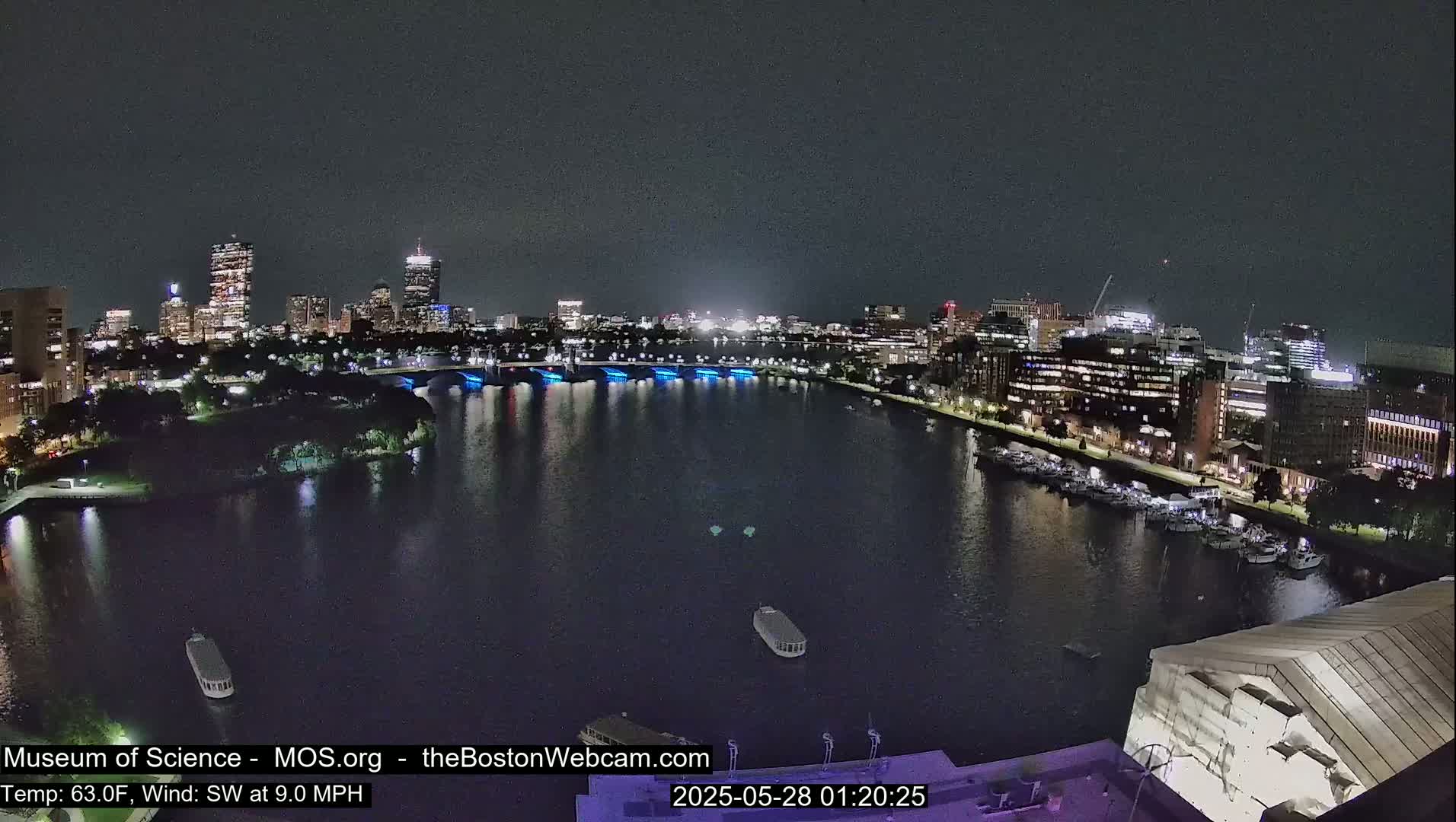 A nighttime view of a city skyline across a calm river, with boats docked along the shore under a clear sky.