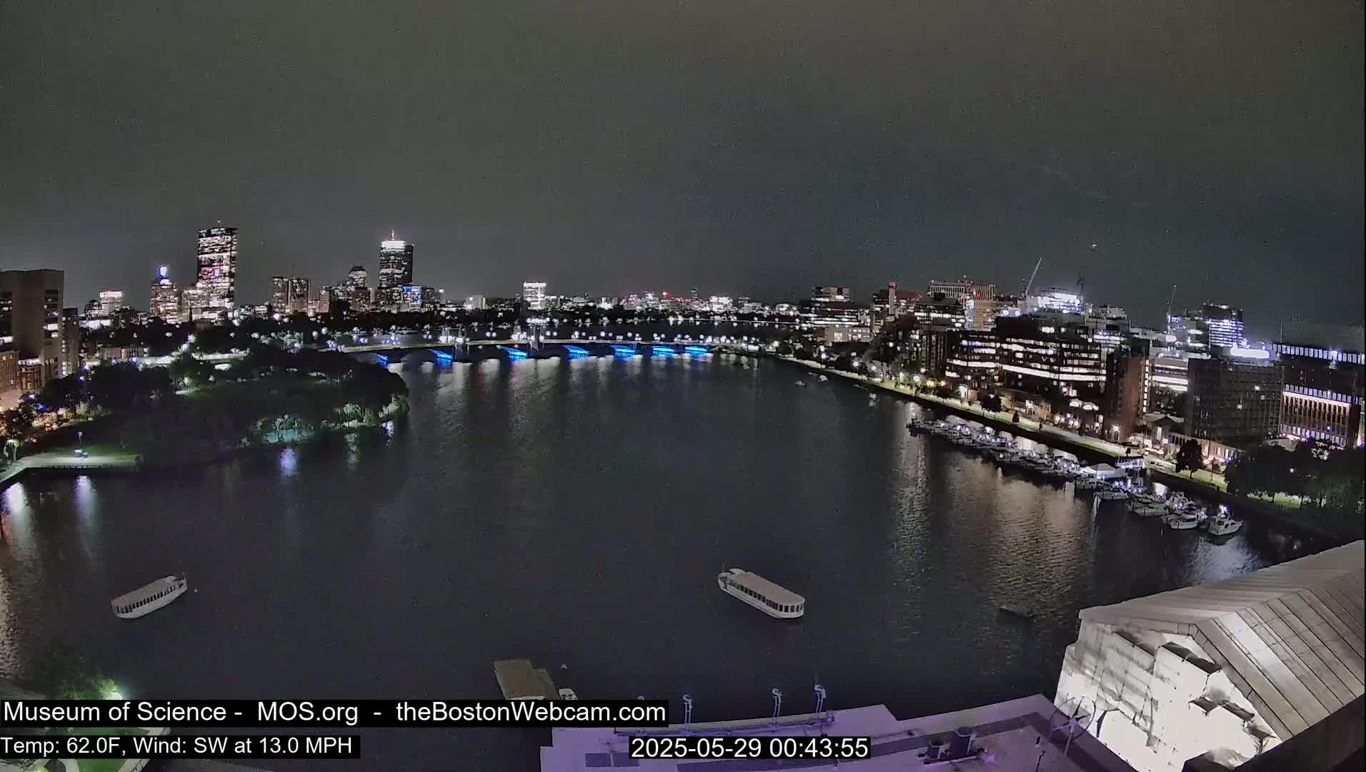 A nighttime view of a city skyline across a calm river, with a southwest wind at 13 mph and a temperature of 62 degrees Fahrenheit.