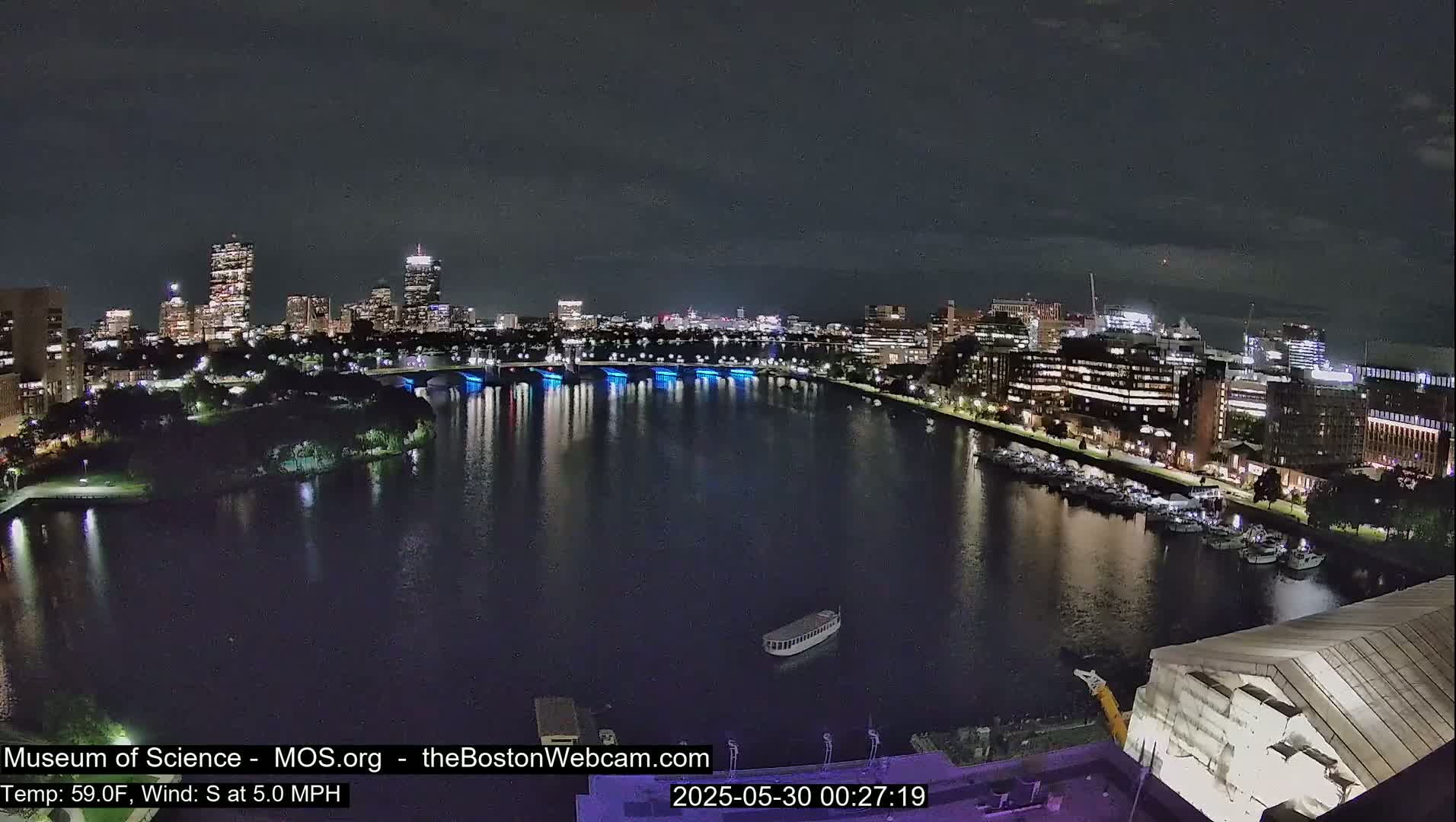 A nighttime view of a city skyline across a calm river, with a gentle south wind and a temperature of 59°F.