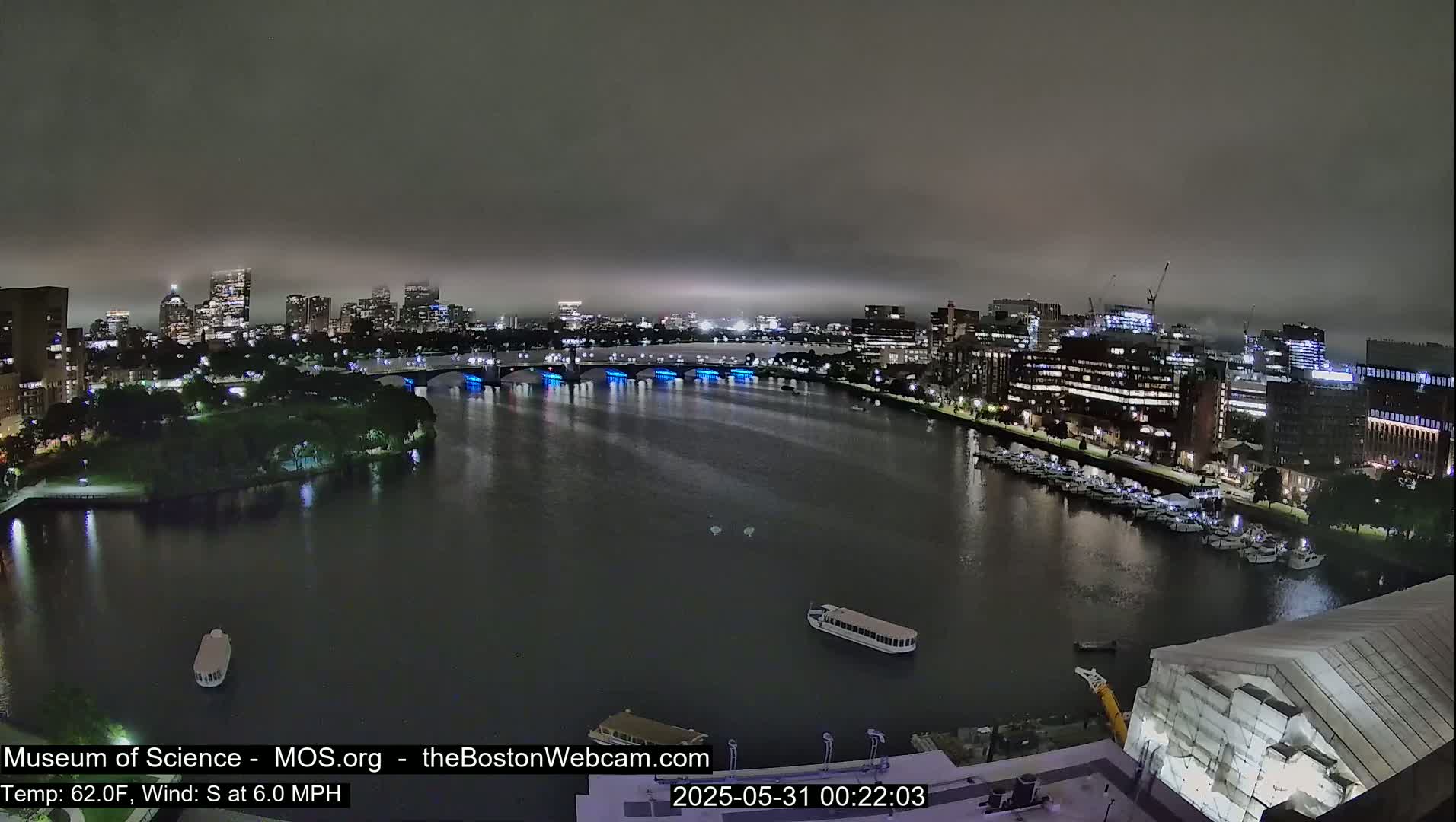 A nighttime view of a city skyline across a river, under a hazy sky, with boats moored along the waterfront.