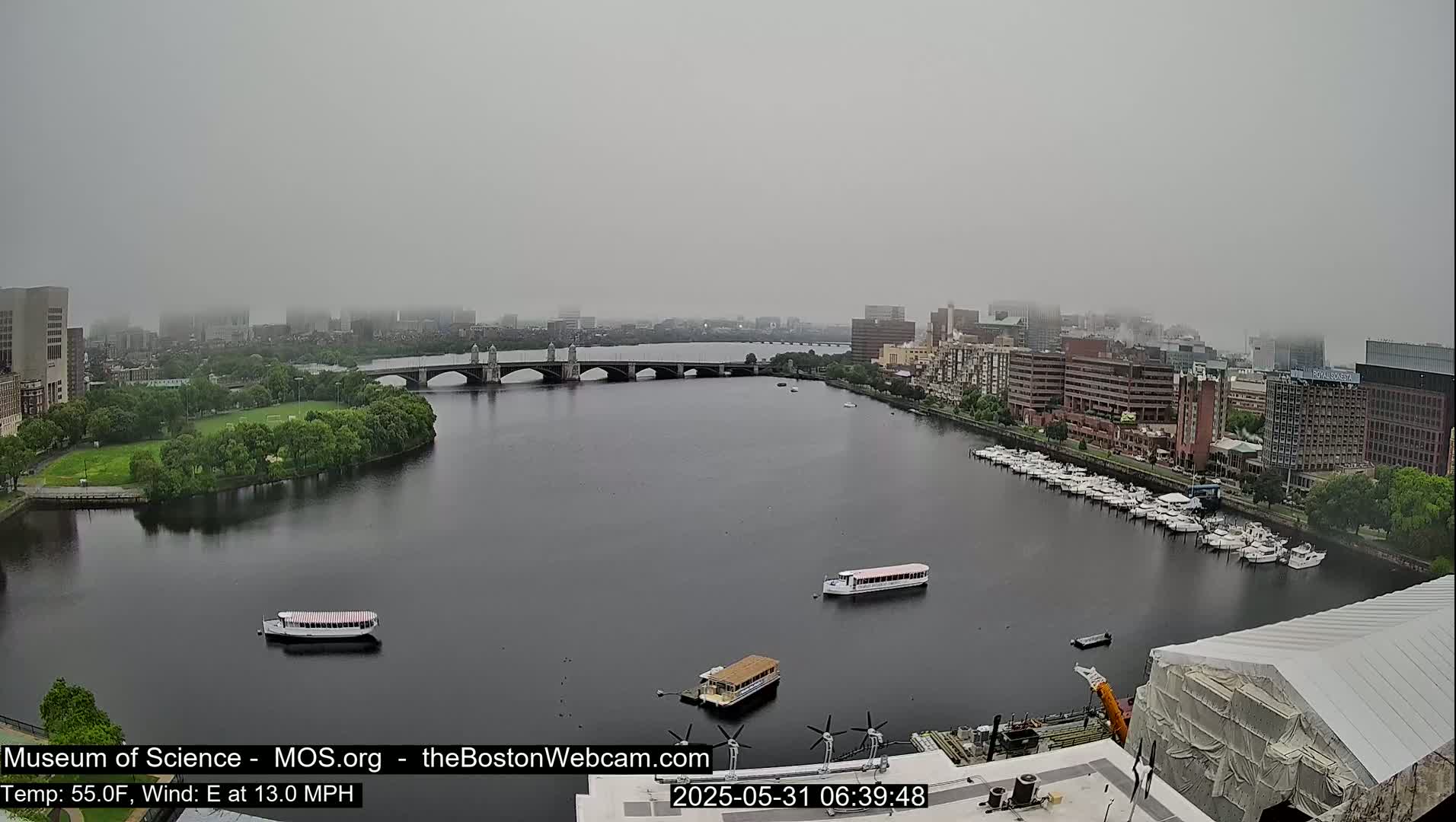 A hazy, overcast view of a river with a bridge, boats, and city buildings in the background.