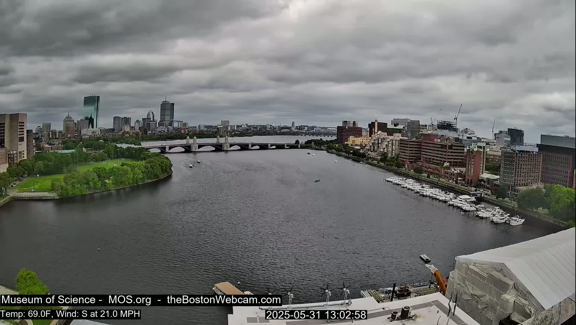 A wide shot of a cityscape under an overcast sky shows a river with a bridge and many docked boats, a green park, and numerous buildings.