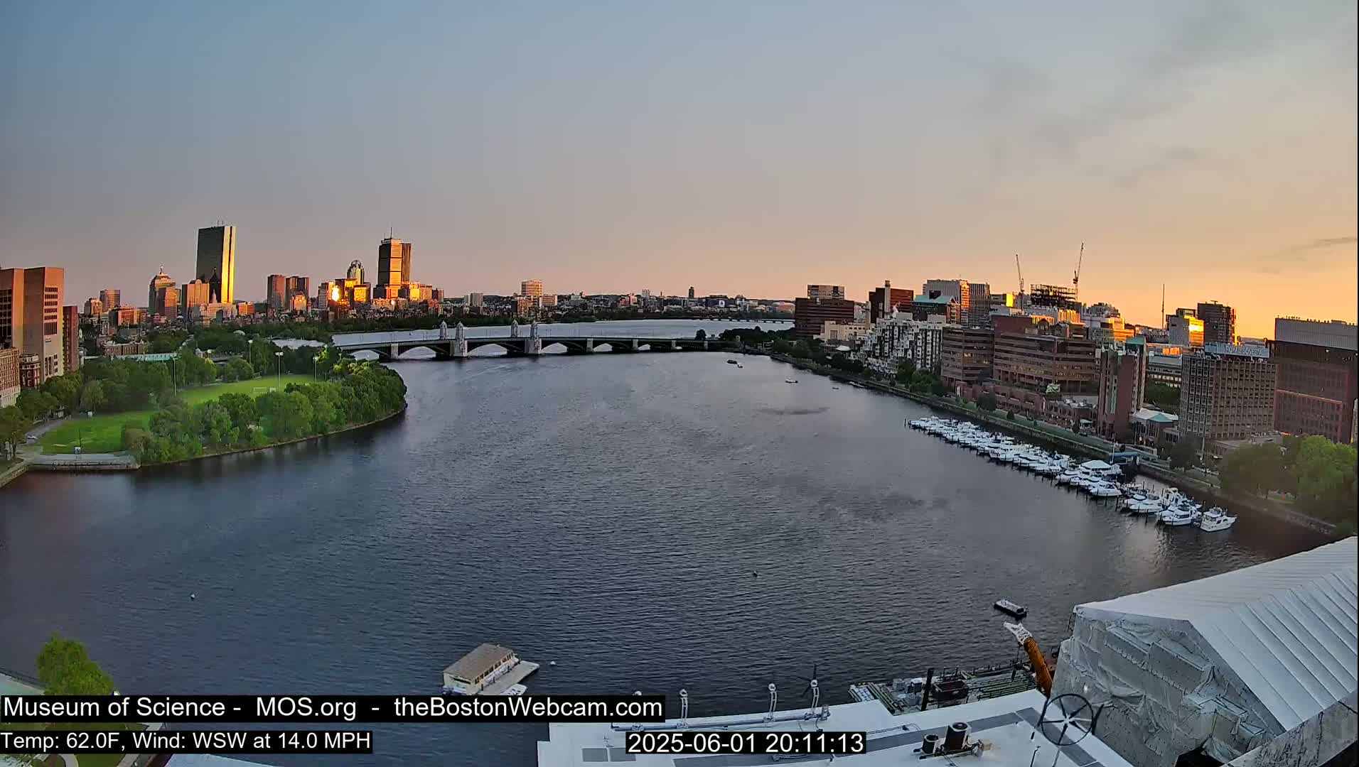 A city skyline at sunset is visible across a river with a bridge, boats docked along the shore, and a park in the foreground, under a mostly clear sky.