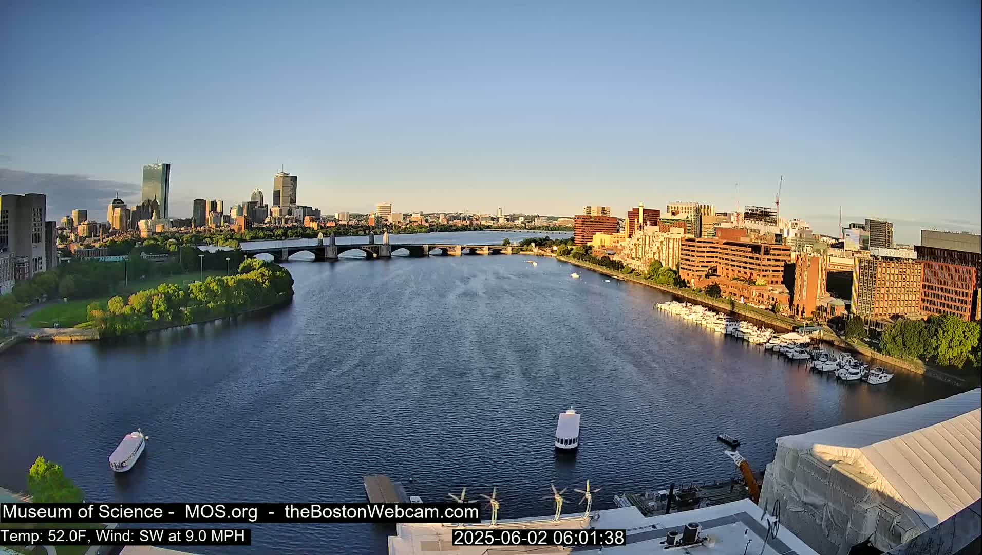A city skyline is visible across a calm river under a clear, bright sky, with boats docked along the riverbank and several bridges spanning the waterway.