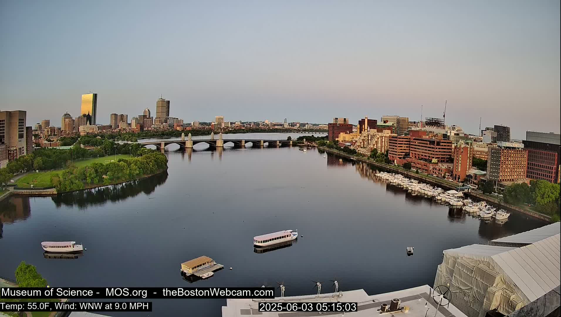 A calm river reflects a city skyline under a clear, light-blue sky at dawn, with several boats moored along the shoreline and a bridge spanning the water.