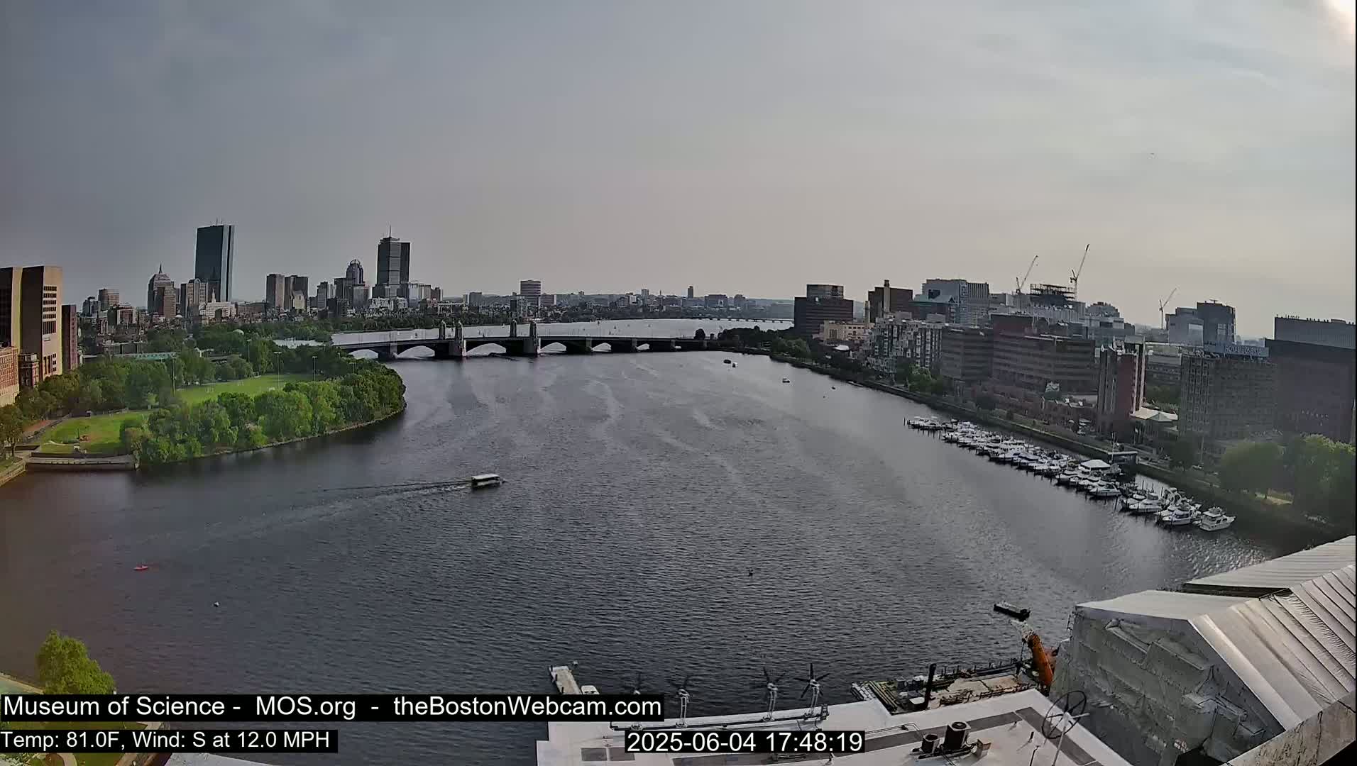 A wide shot shows a city skyline across a river with a bridge, boats docked on the opposite shore, and a partly cloudy sky.