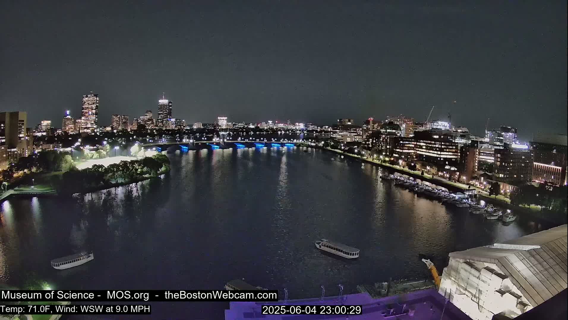 A nighttime view of a city skyline across a dark river, with boats docked along the shore and a bridge lit with blue lights, under a clear sky.