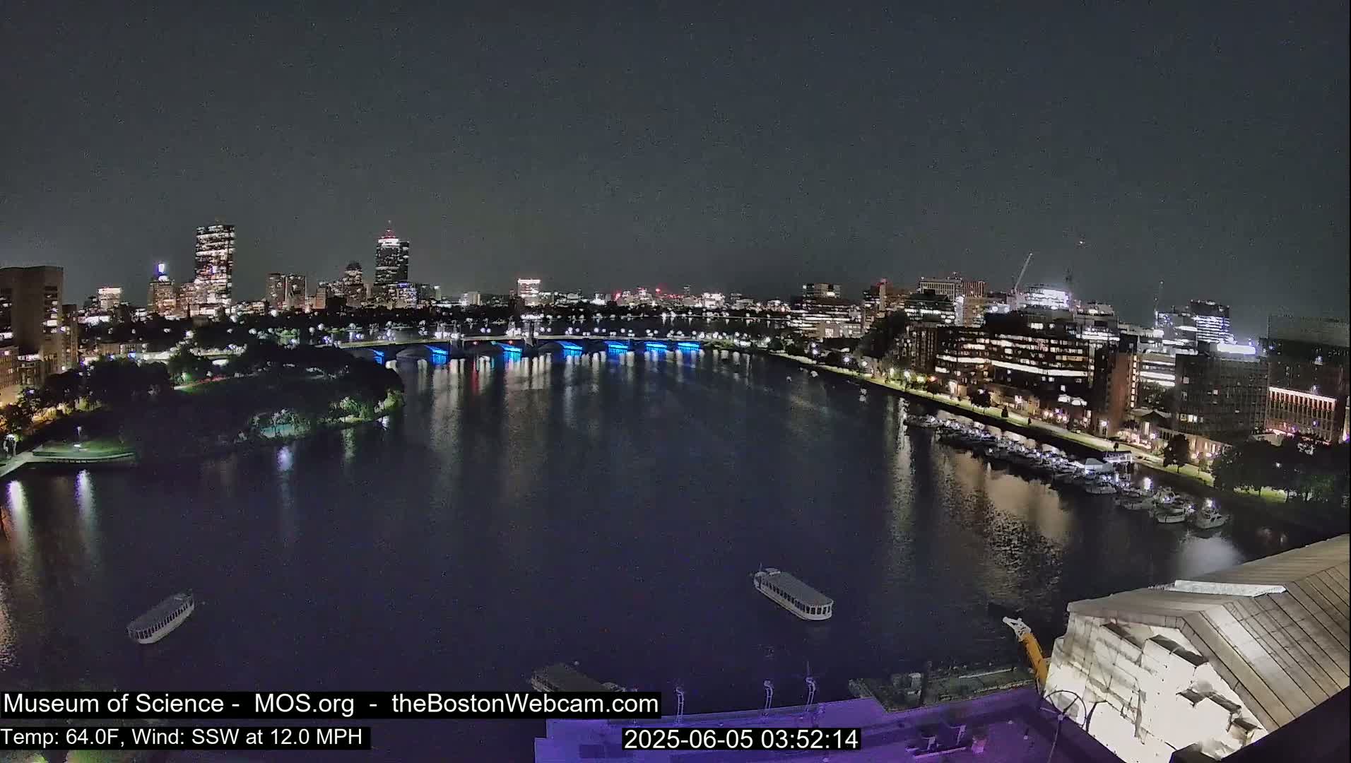 A nighttime view of a city skyline across a calm river, with a gentle south-southwesterly wind at 12 mph and a temperature of 64°F.