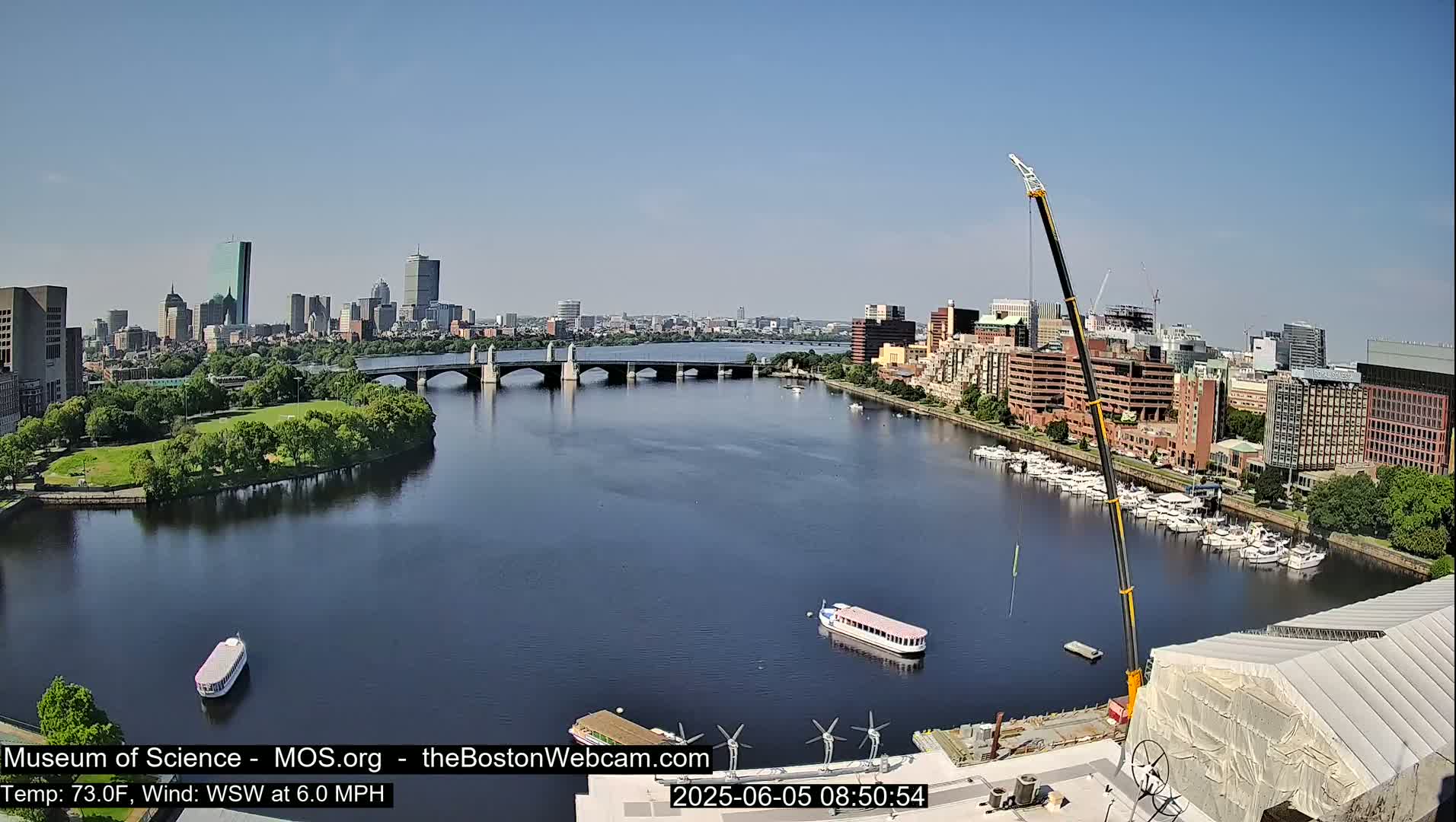 A sunny cityscape with a river, bridge, boats, and a construction crane is visible on a clear day.