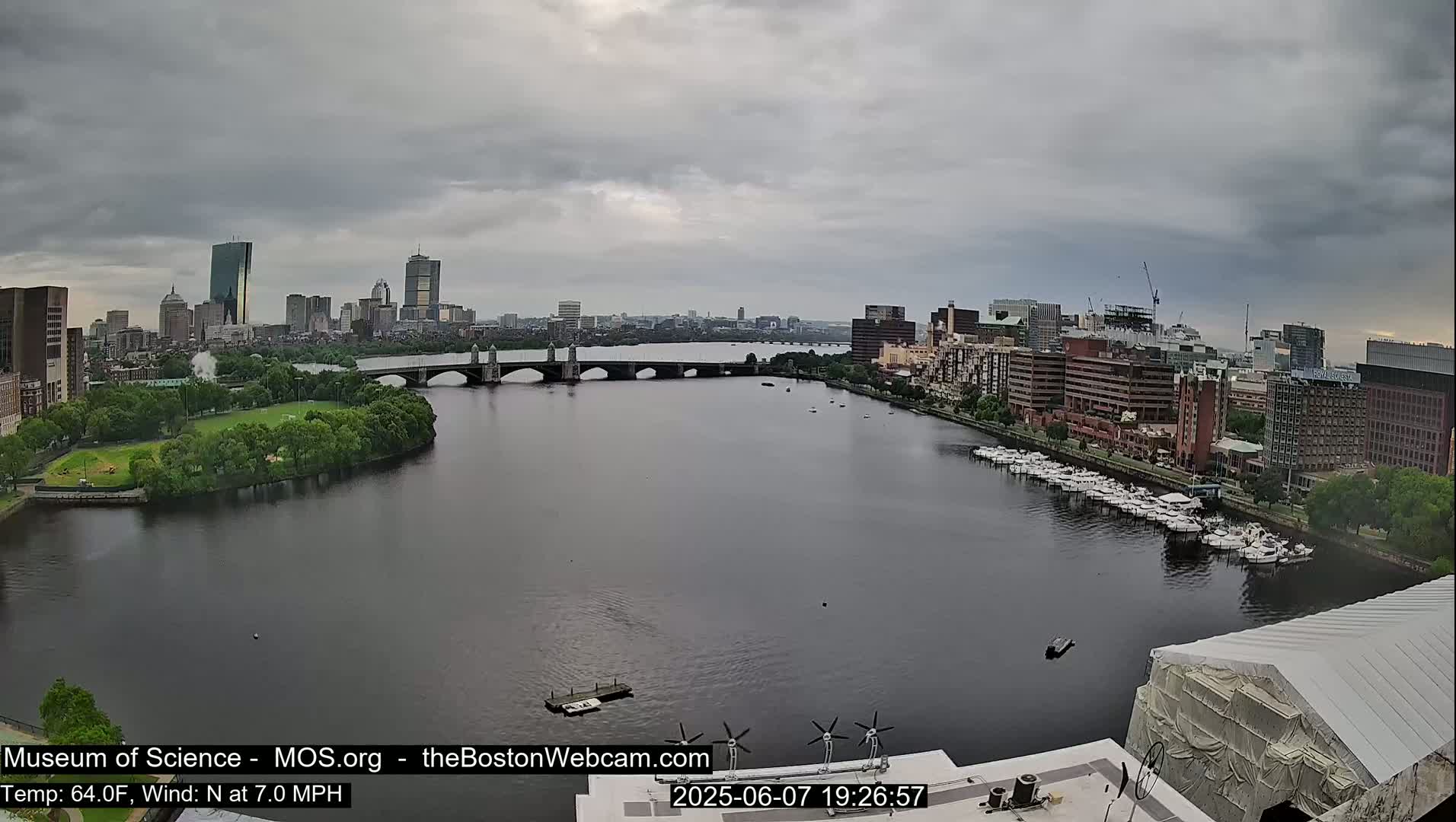 A wide shot shows a city skyline across a river under an overcast sky, with a bridge and docked boats visible.