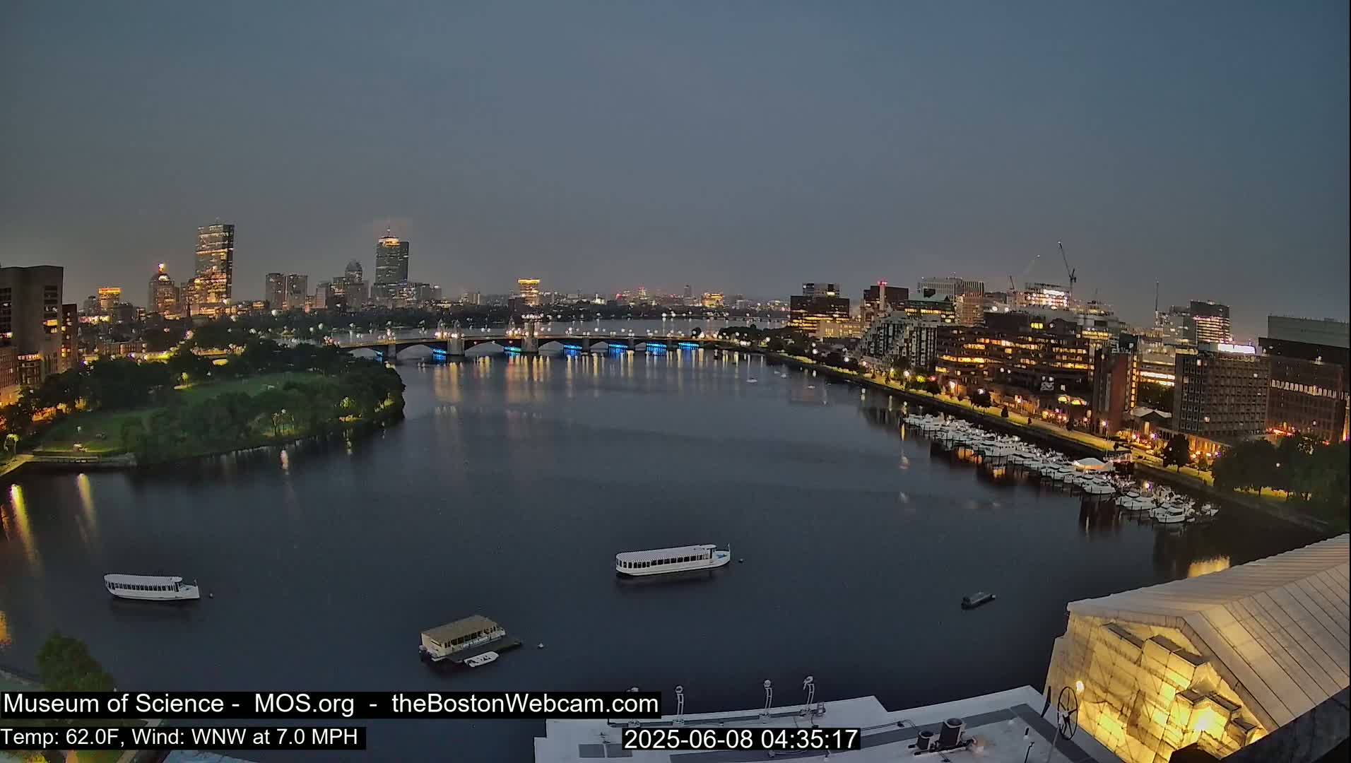 A nighttime view of a city skyline across a calm river, with several boats docked and a few on the water, under a dusky sky.