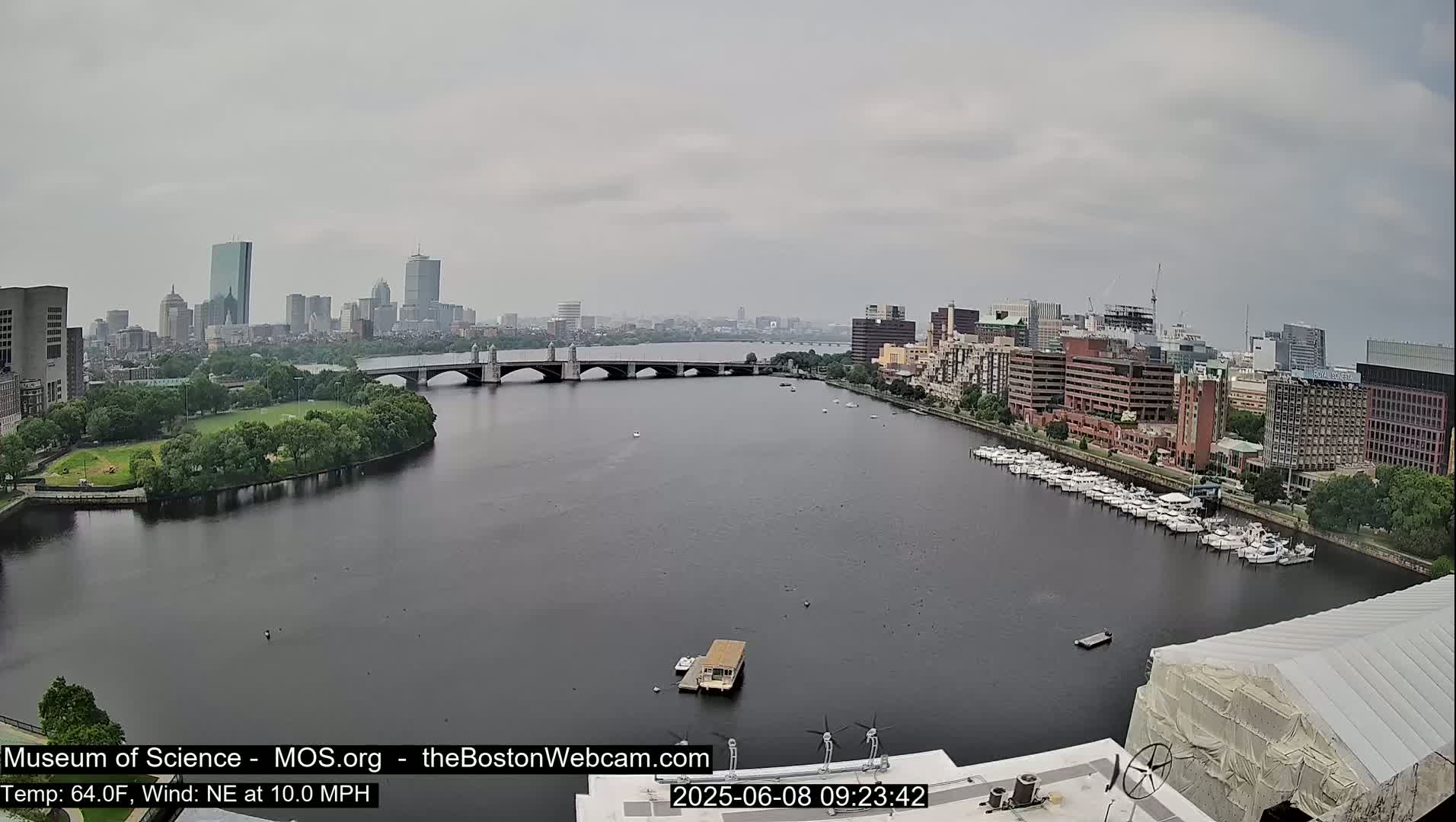 A city skyline is visible across a wide river on an overcast day, with a bridge spanning the waterway and numerous boats moored along the shore.