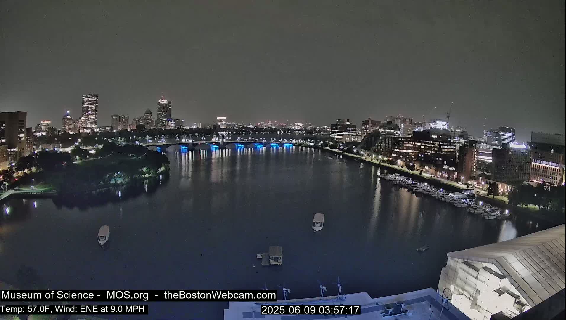 A nighttime view of a city skyline across a calm river, with a bridge illuminated in blue, under a dark sky.