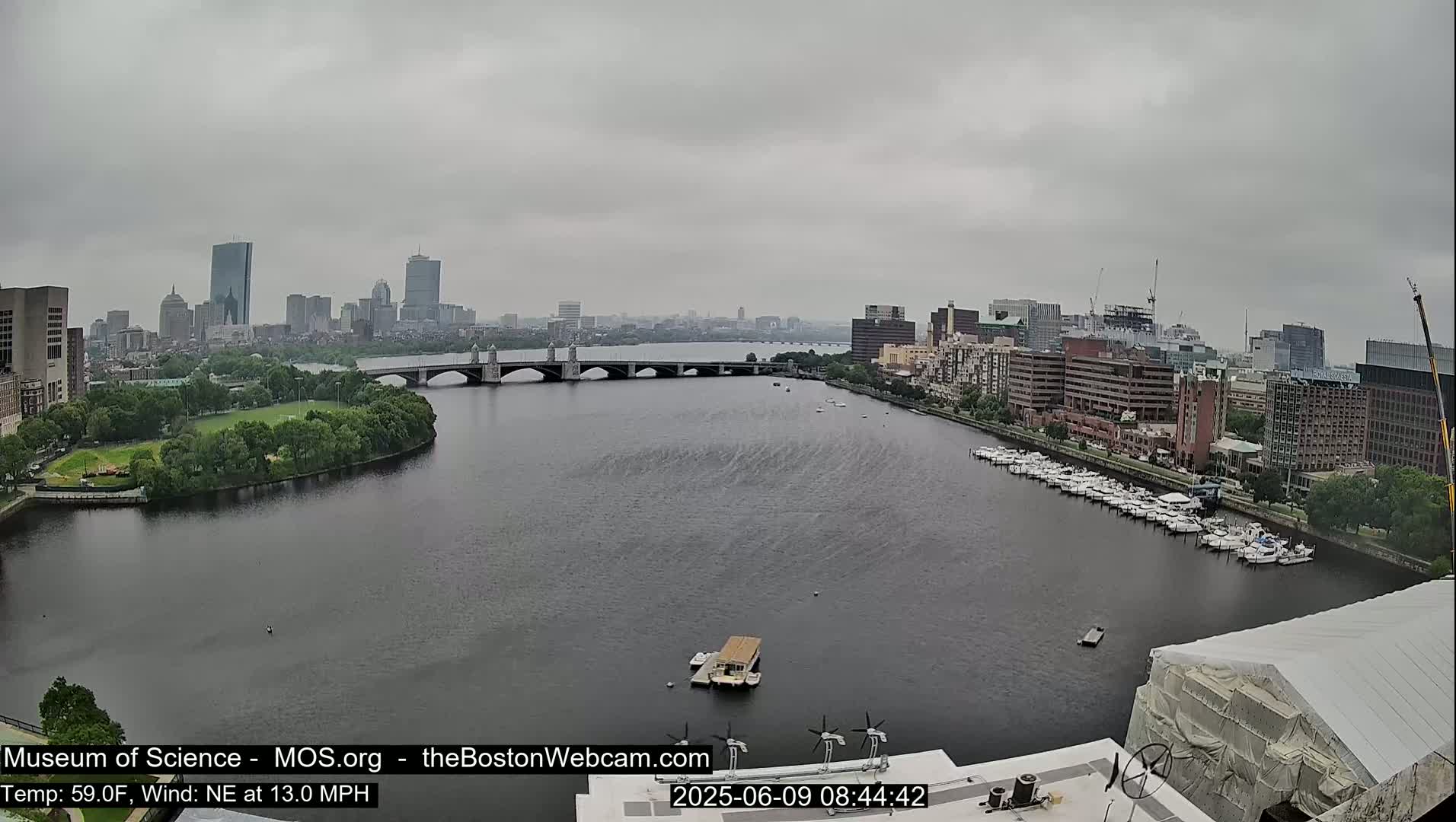 A wide shot of a city skyline across a river on an overcast day, with boats docked along the shore and a bridge in the middle ground.