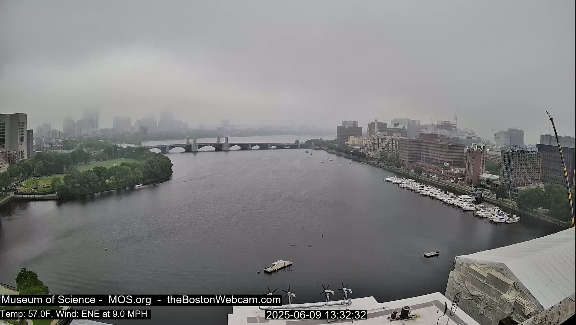 A hazy, overcast day shows a river with a bridge and docked boats, and a city skyline in the distance.