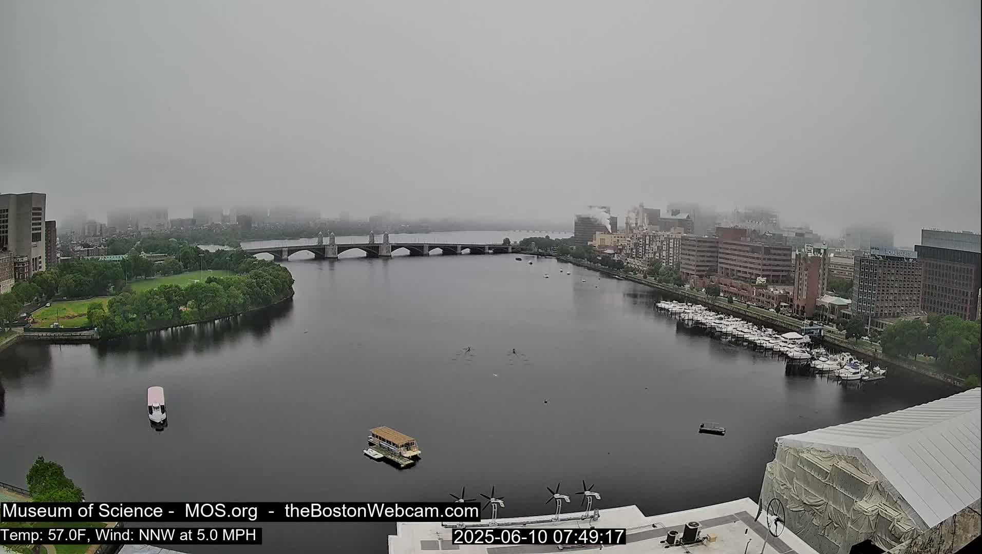 A foggy cityscape is viewed from a high vantage point, showing a river with boats, a bridge, and buildings on either side.