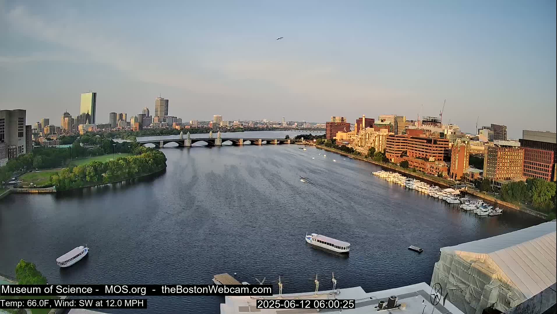 A city skyline is visible across a river with a bridge, boats, and docked yachts under a clear, mostly sunny sky.