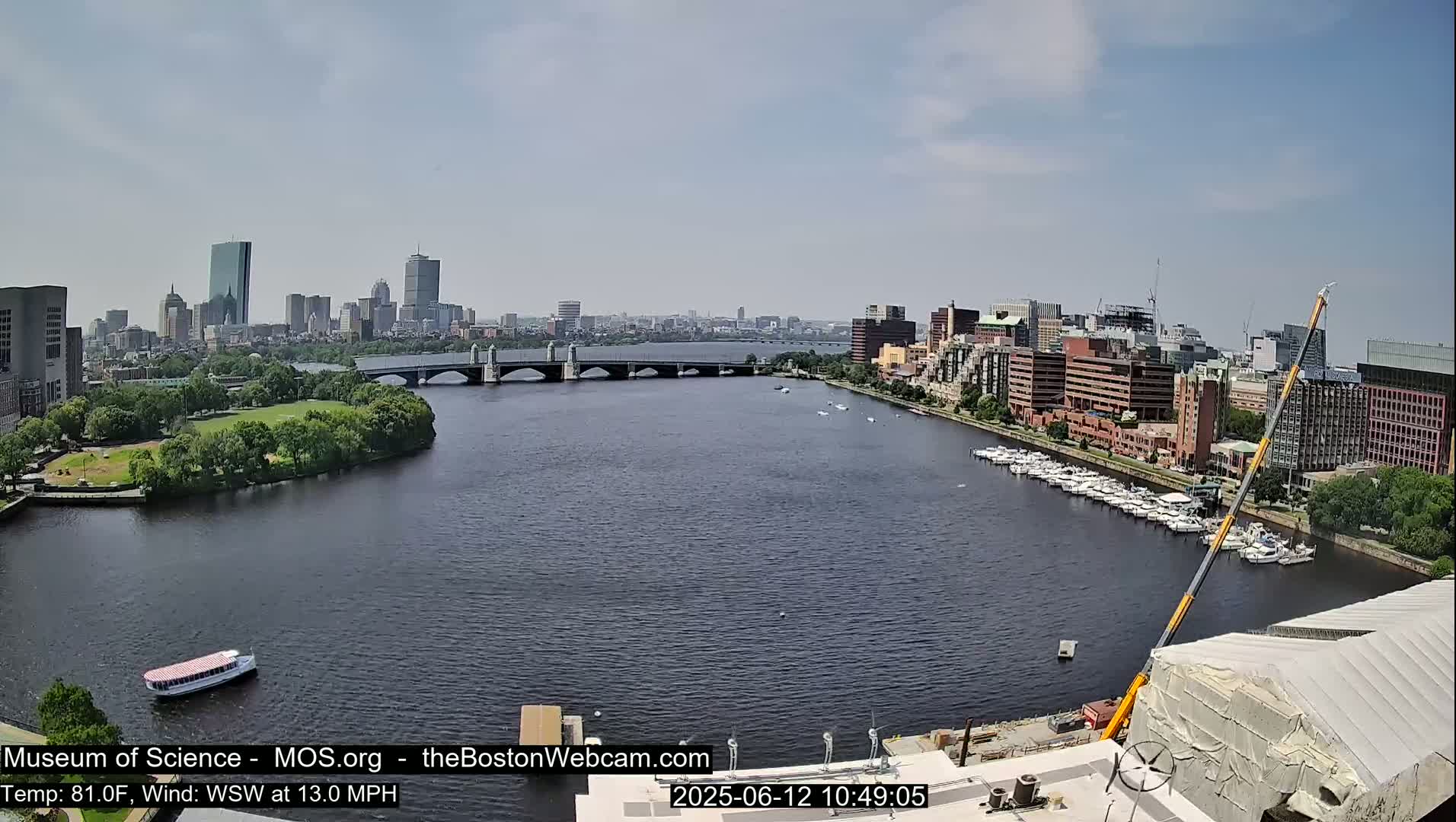A long shot of a city skyline across a river on a partly sunny day with a moderate wind, showing a bridge, boats, and a crane near a building under construction.