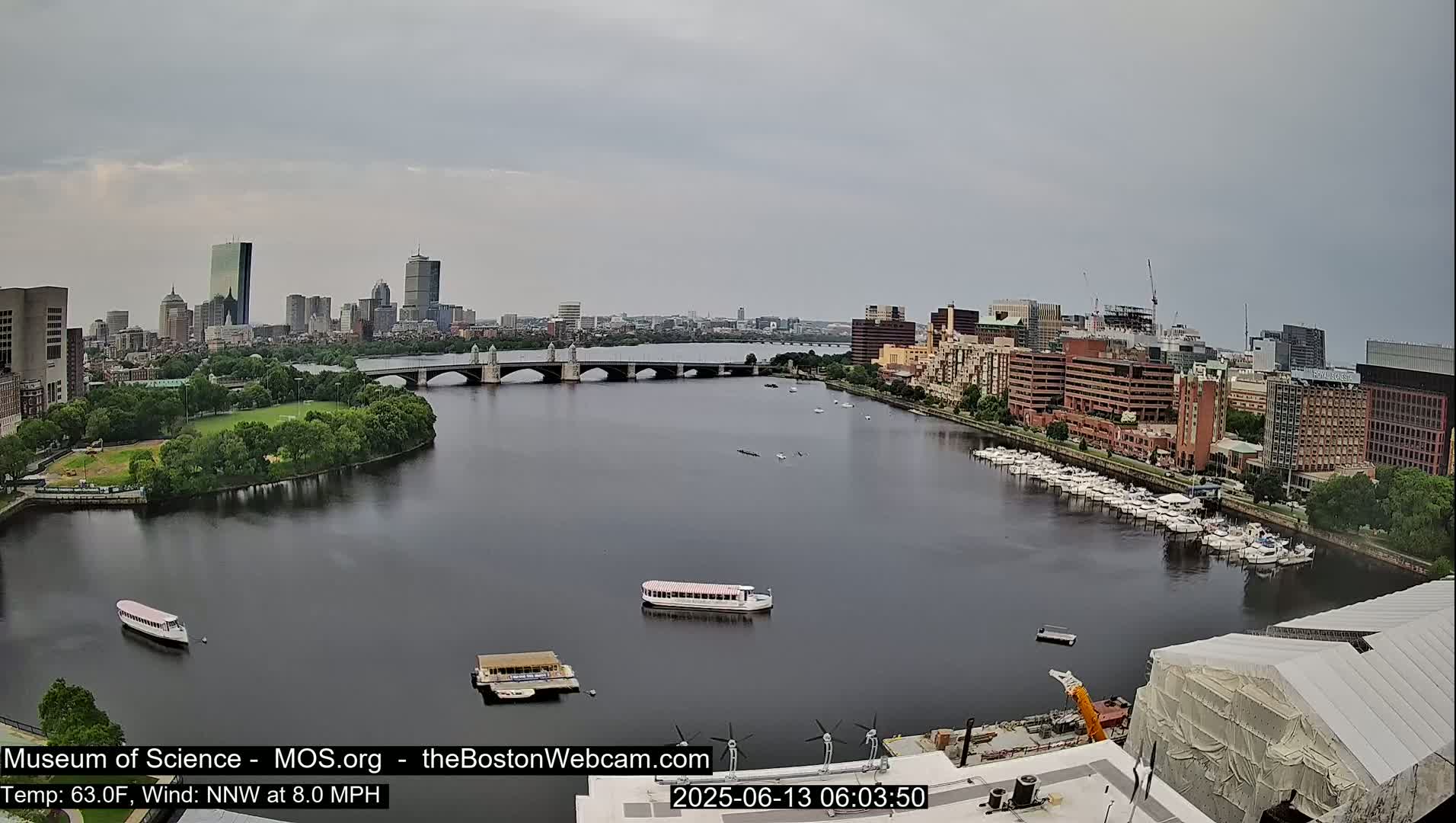 A wide shot of a city skyline across a calm river, under an overcast sky, shows a bridge, several boats, and a marina.