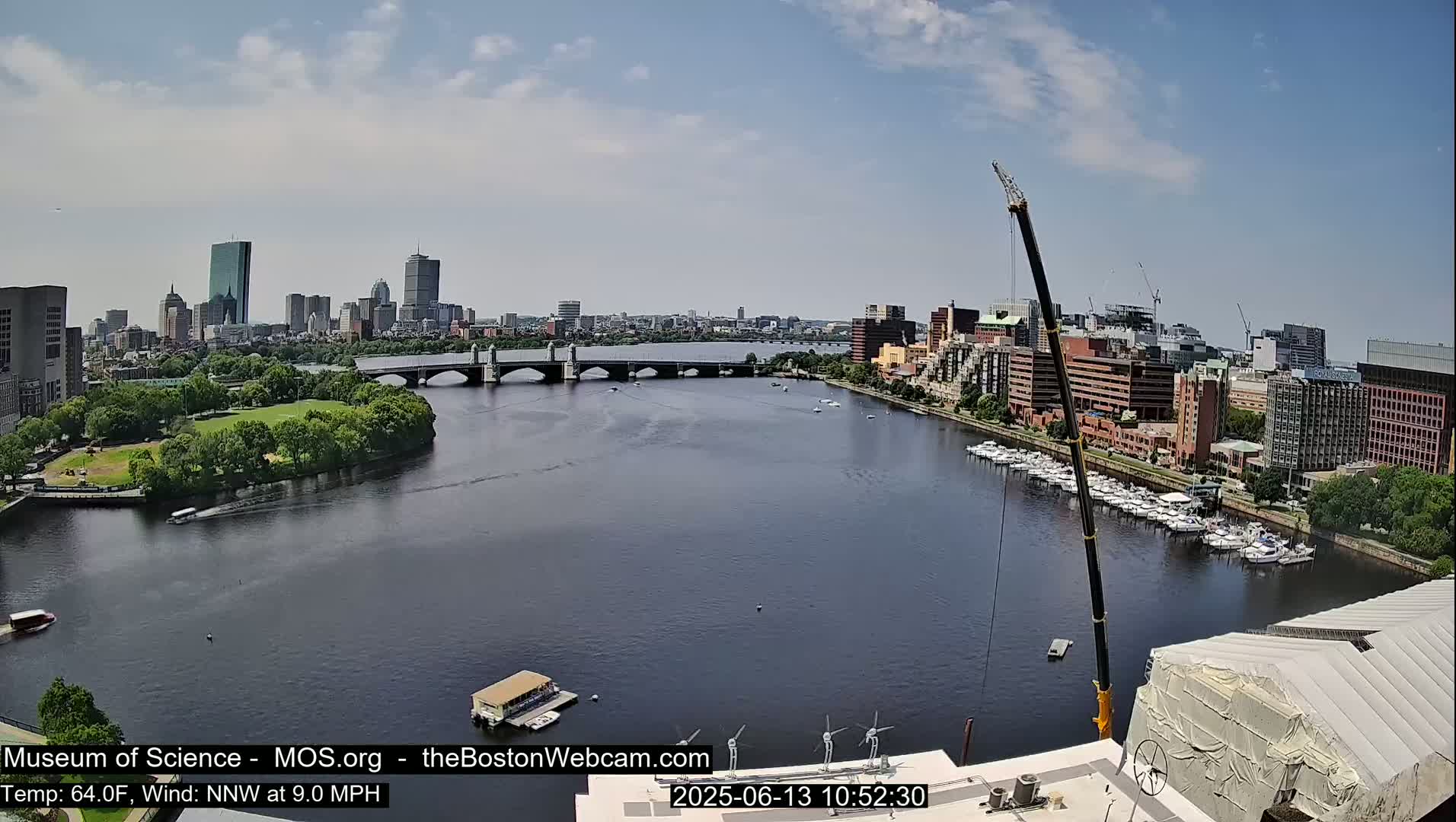 A city skyline with a river, bridge, and docked boats is visible under sunny skies, with a large crane present near the foreground.