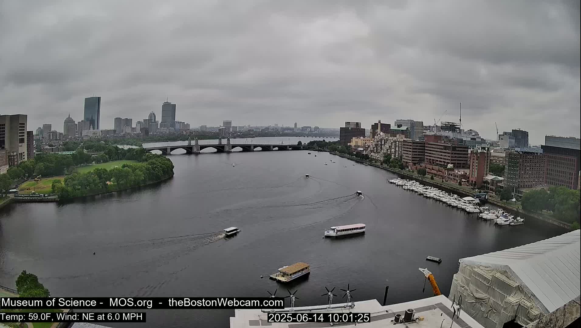 A river flows through a city under an overcast sky, with several boats on the water and a bridge spanning the waterway.