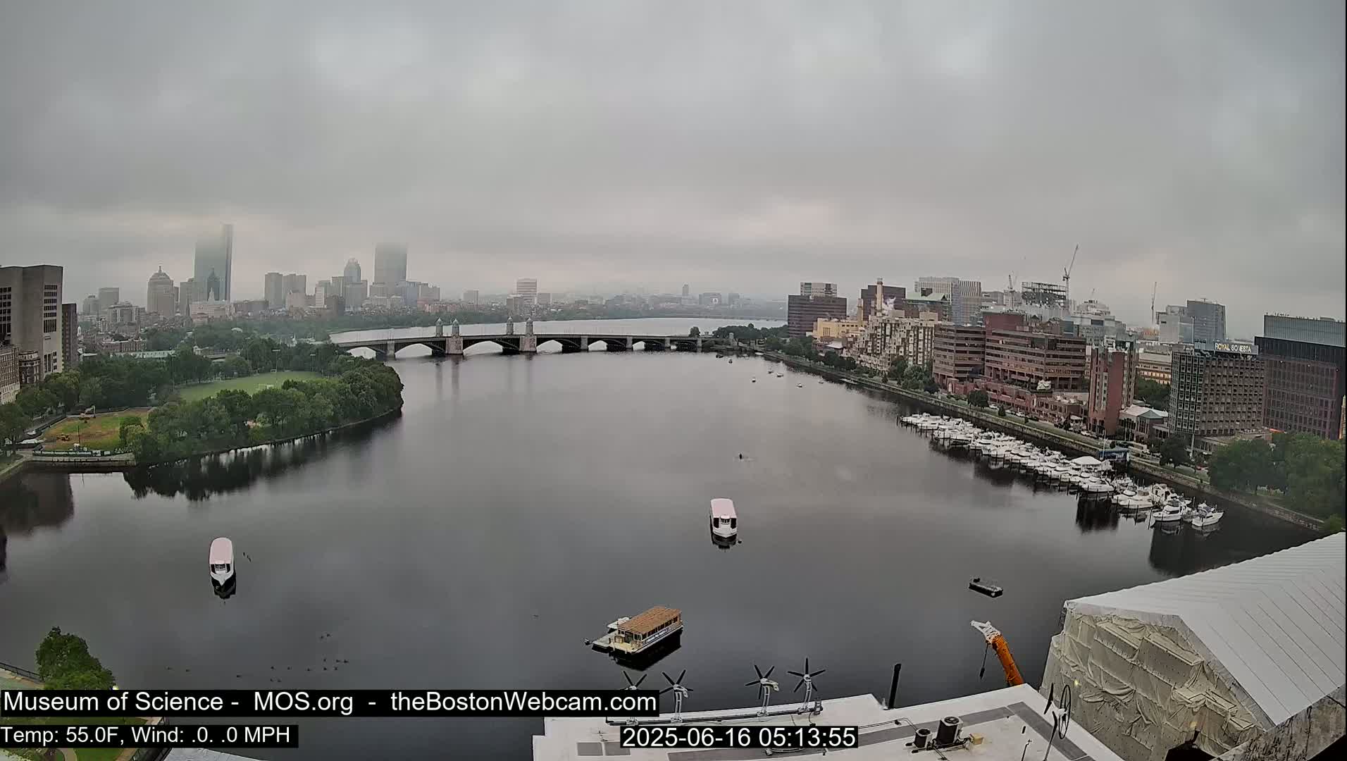 A city skyline is visible across a calm river on an overcast day, with a bridge and boats present.