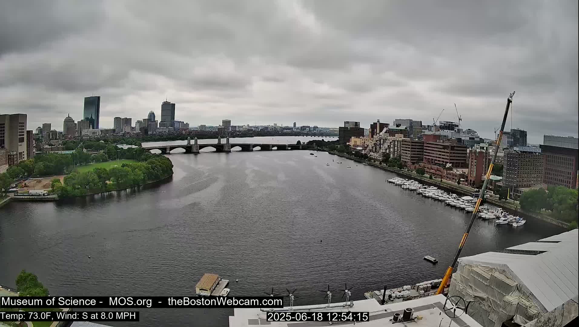 A river flows through a city under an overcast sky, with a bridge spanning the river and numerous boats docked along the shore.