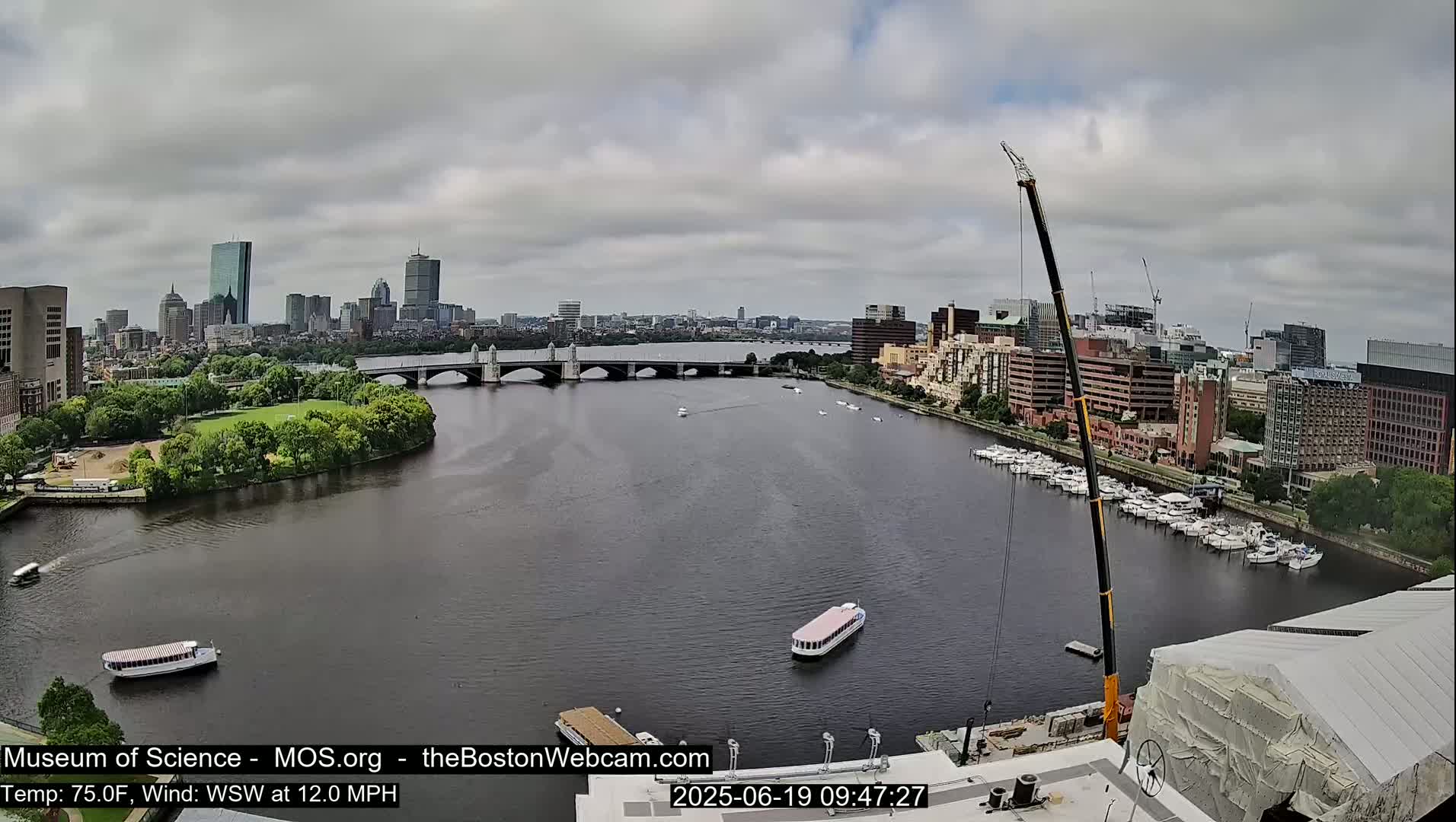 A wide shot shows a cityscape across a river under an overcast sky, with a bridge, boats, and a construction crane visible.