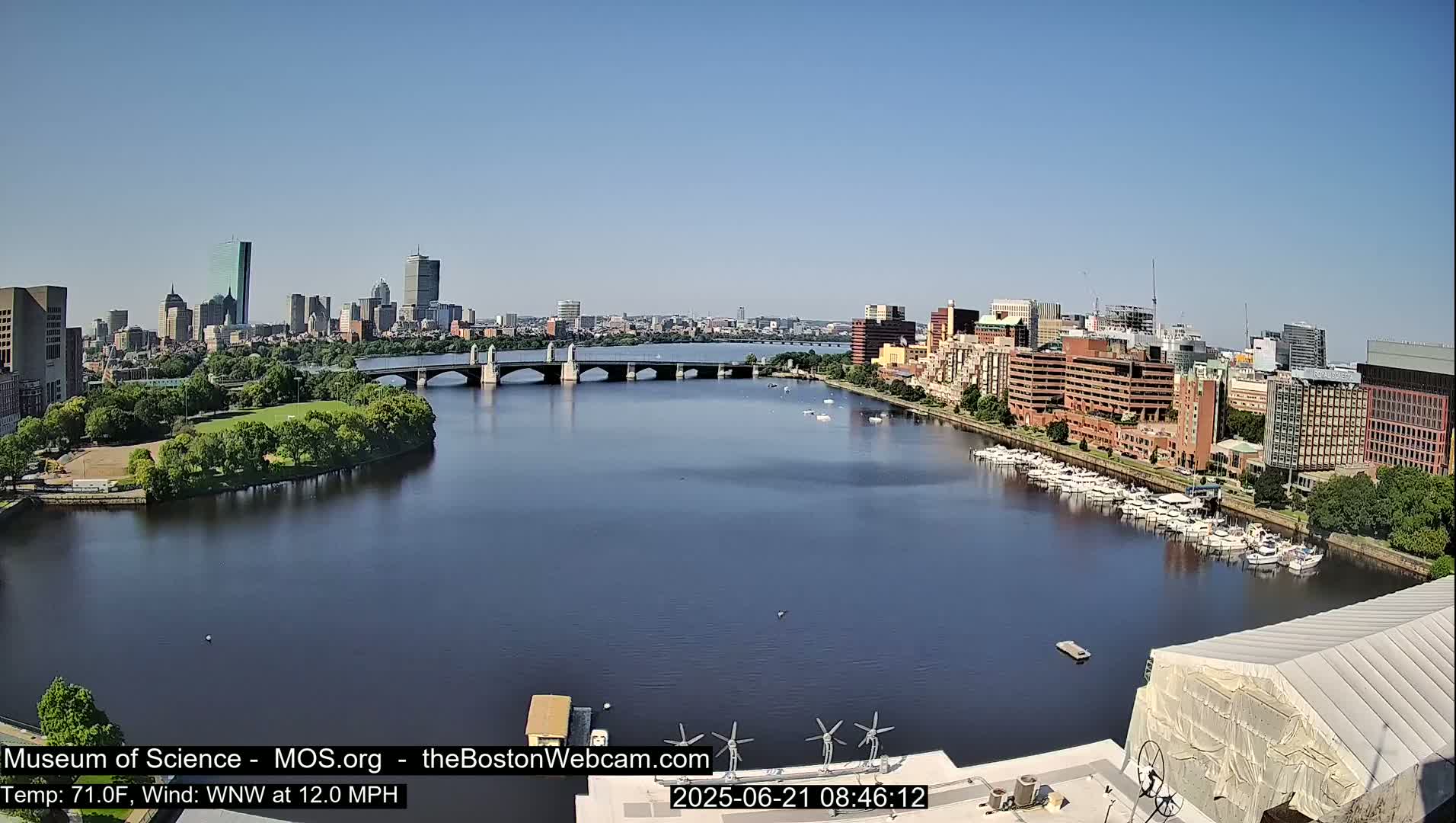 A long shot of a city skyline across a river on a sunny day, with a bridge spanning the river and numerous boats docked along the shore.