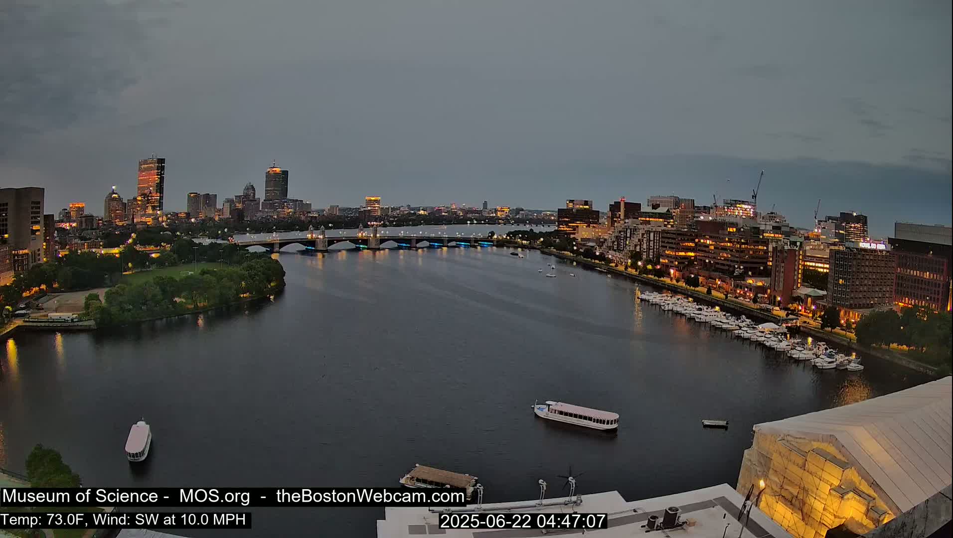 A nighttime cityscape with a river, bridge, and many boats, under a cloudy sky.