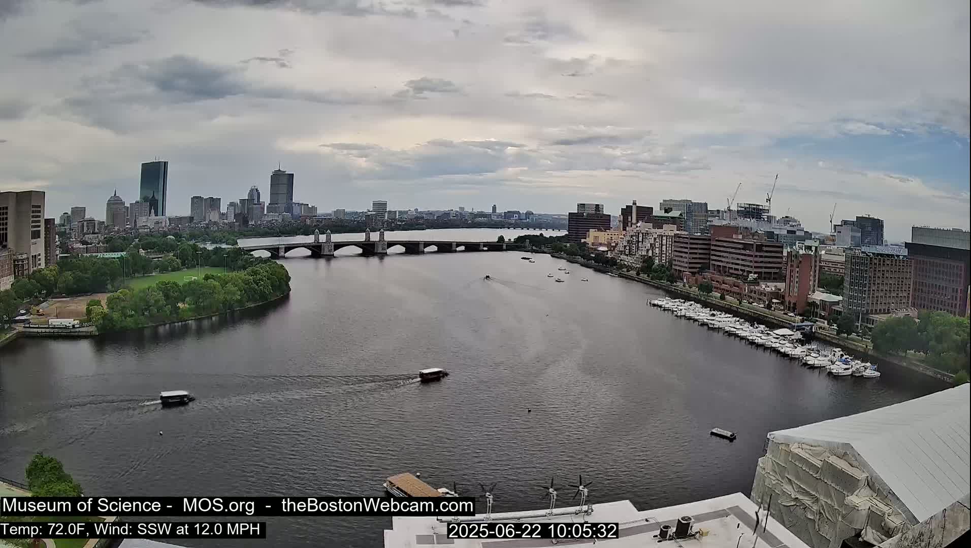 A city skyline is visible across a wide river with a bridge and docked boats under an overcast sky.