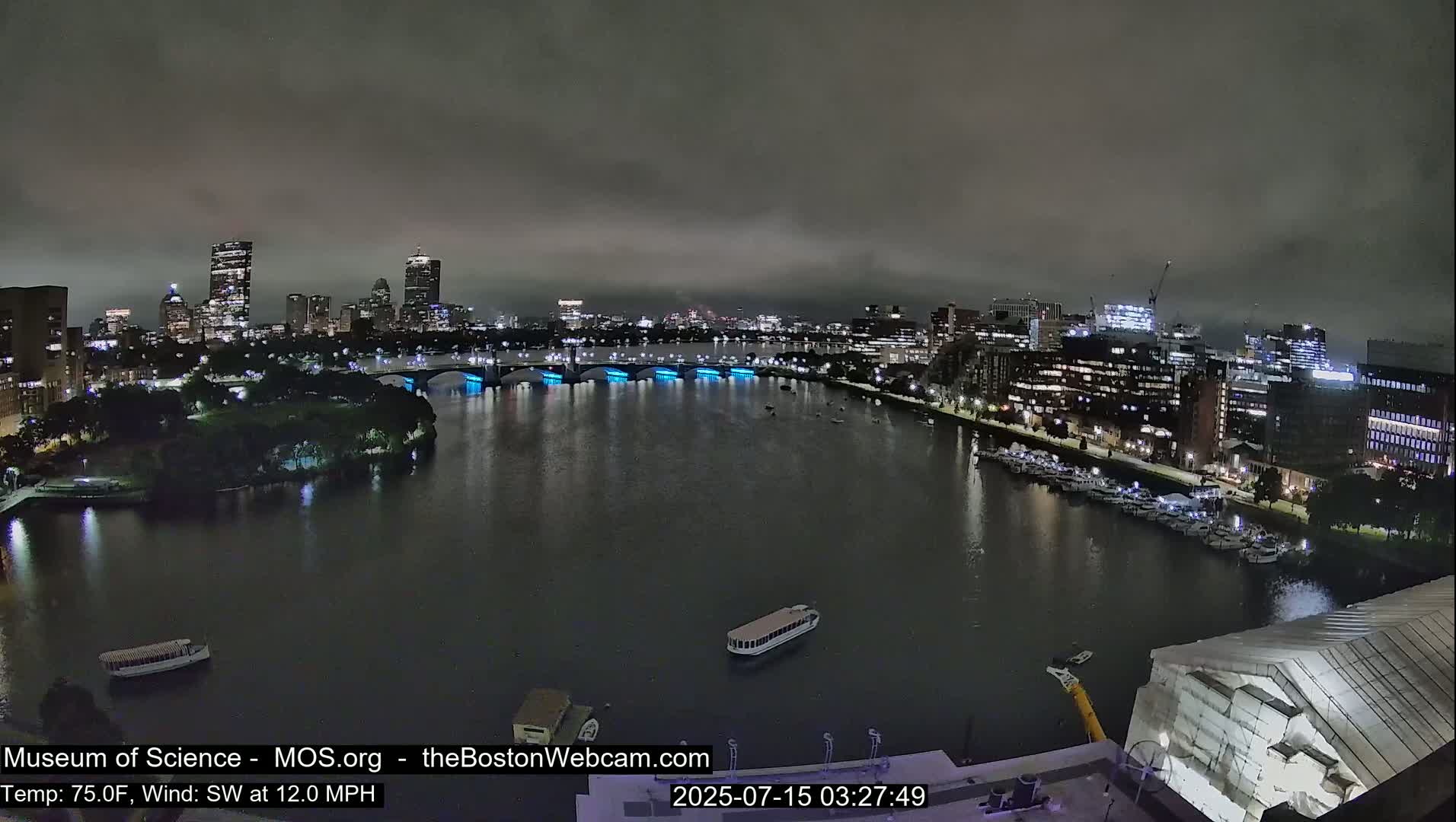 A nighttime view of a city skyline across a dark river under an overcast sky, with a boat on the water and lights illuminating a bridge.