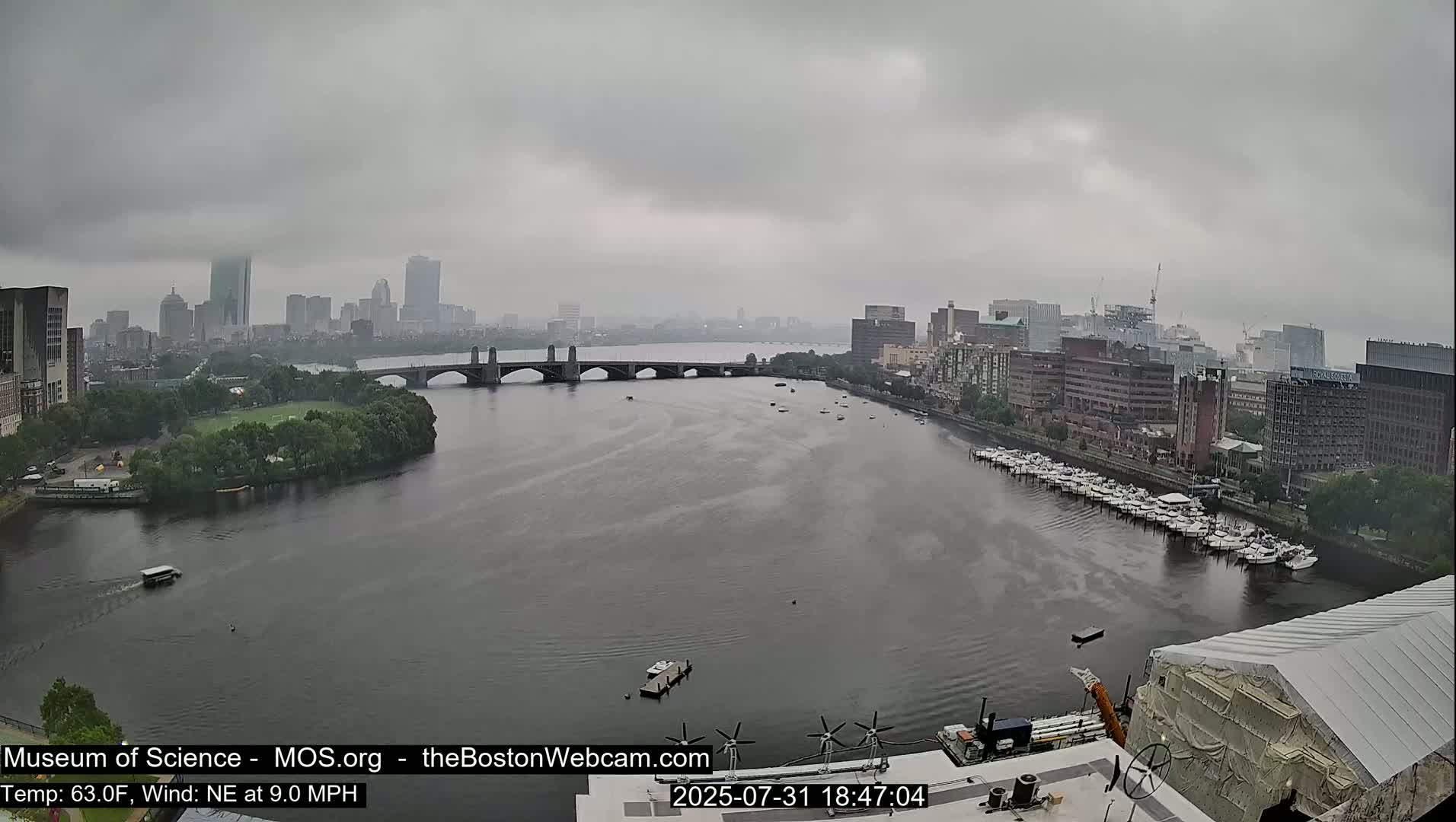 A wide shot of a city skyline across a river under an overcast sky, showing a bridge, boats docked along the shore, and some construction.
