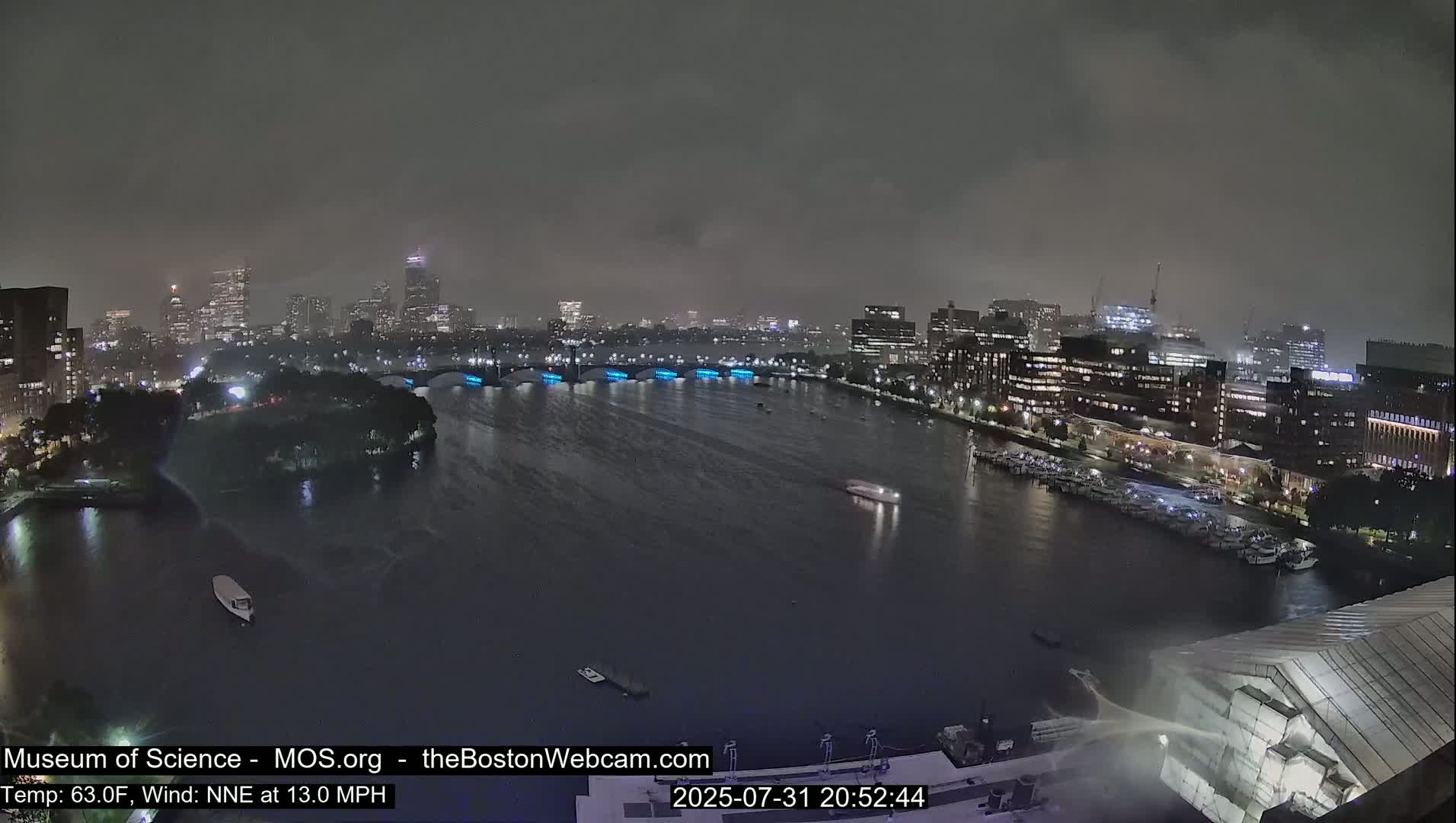 A wide shot of a city skyline across a river under an overcast sky, showing a bridge, boats docked along the shore, and some construction.