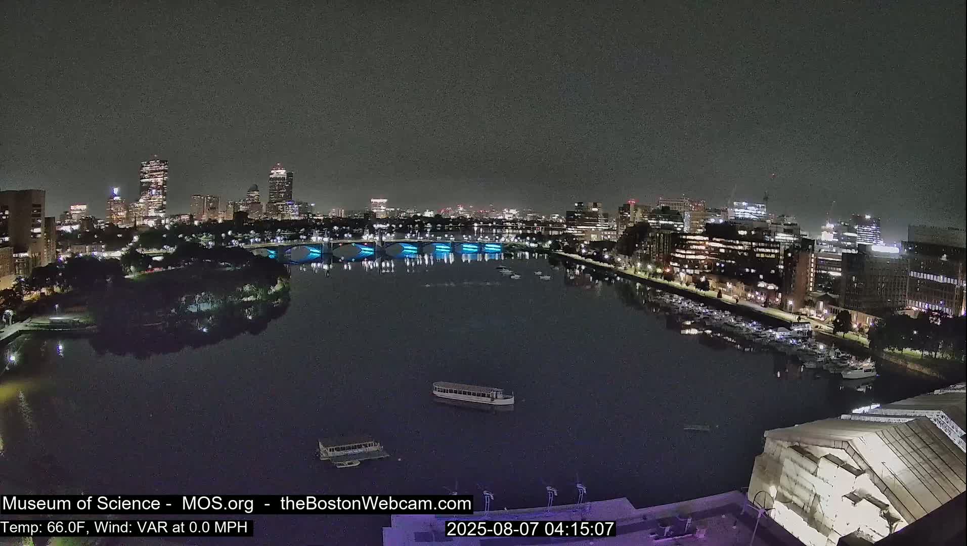 A nighttime view of a city skyline across a calm river, with several boats on the water and lights illuminating a bridge.