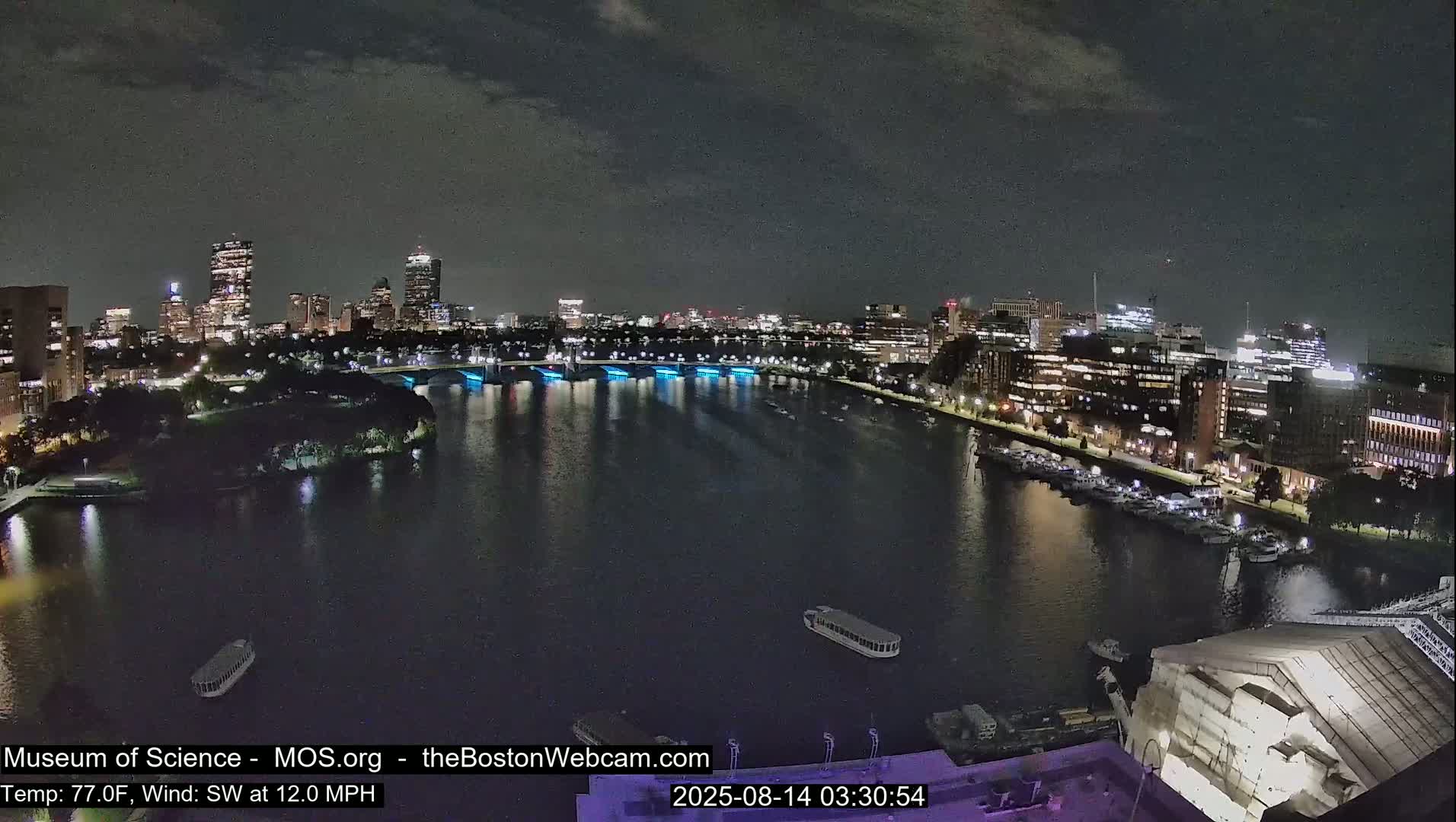 A nighttime view of a city skyline across a river with boats, under a partly cloudy sky, with a 77°F temperature and a 12 mph southwest wind.