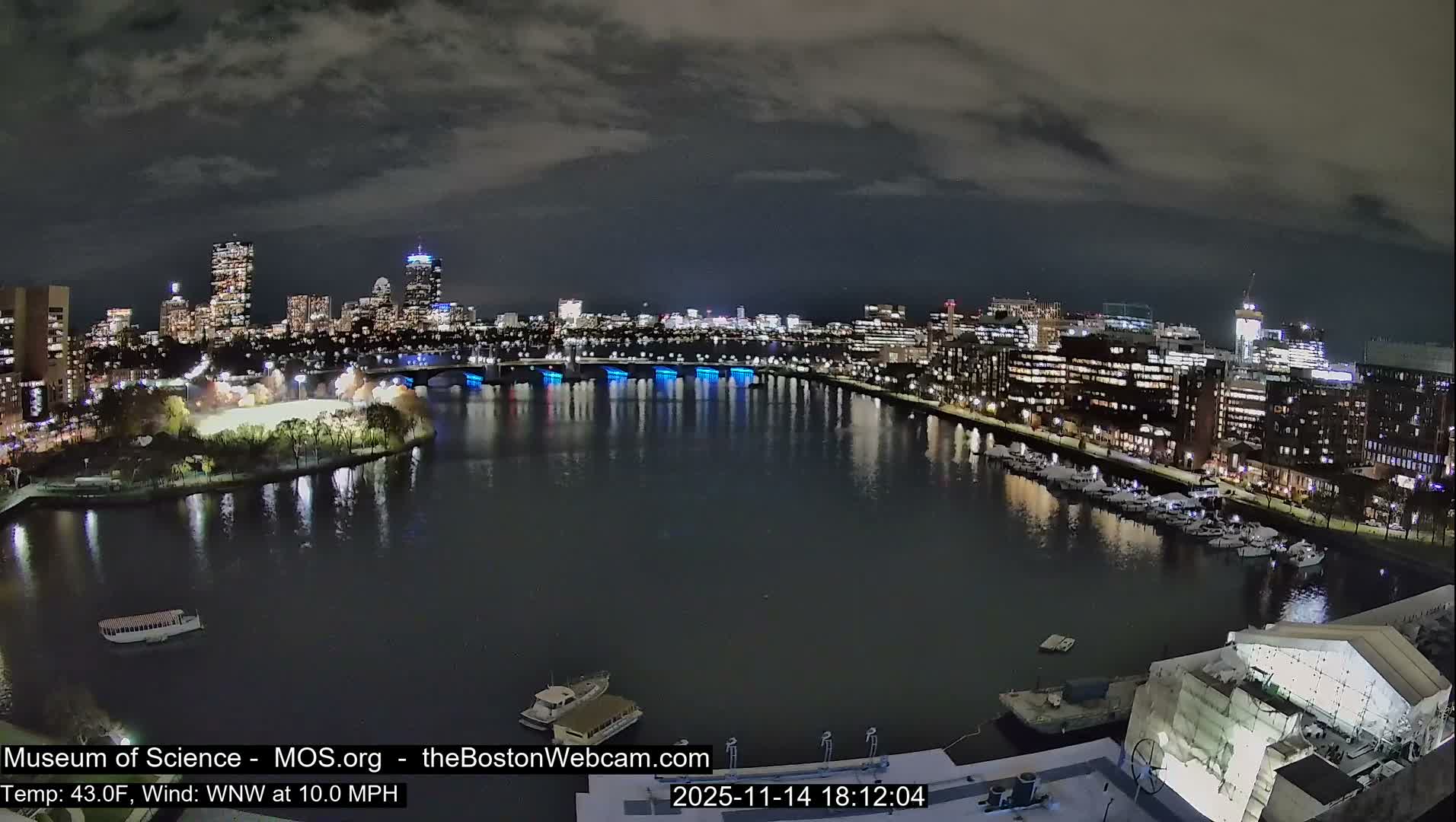 A vibrant city skyline with numerous lit buildings and a blue-lit bridge is reflected in a dark river, featuring several boats, under a cloudy night sky with a temperature of 43.0F and a WNW wind at 10.0 MPH.