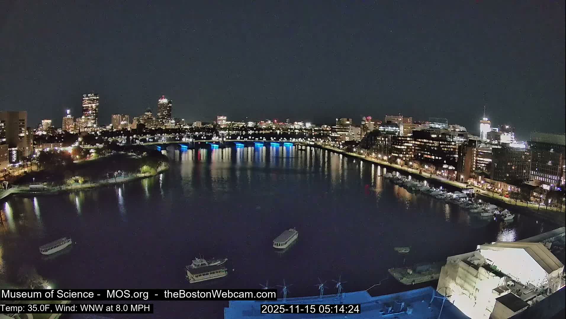 A wide-angle nighttime view shows a clear city skyline with numerous illuminated buildings and a bright blue-lit bridge reflecting in a dark river where several boats float or are docked, all under clear skies at 35.0°F with a WNW wind at 8.0 MPH.