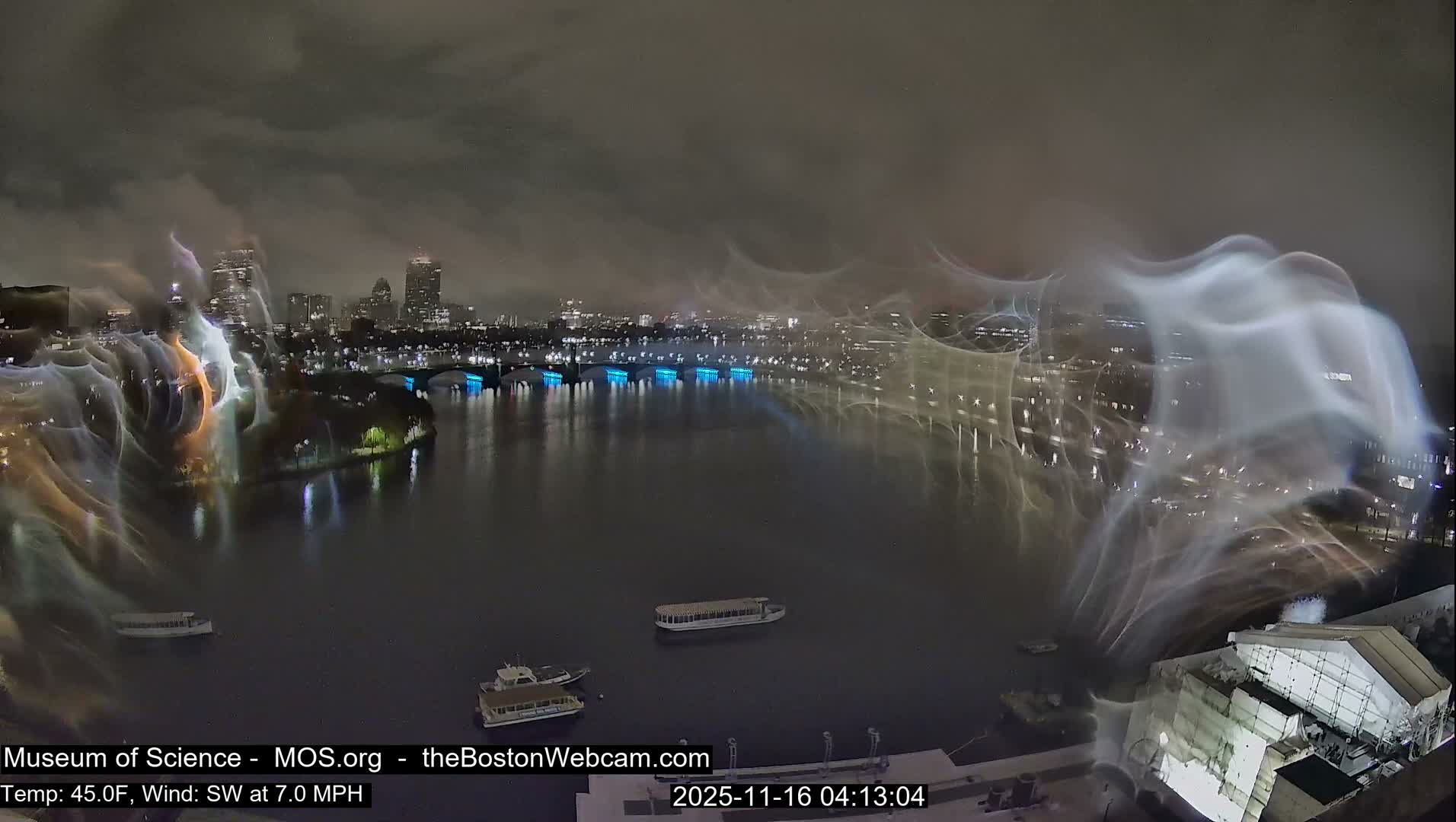 Under an overcast night sky, a city skyline with illuminated buildings and a blue-lit bridge reflects on a dark body of water where several boats float, all heavily obscured by prominent, swirling light streaks across the camera lens, suggesting damp or rainy conditions.