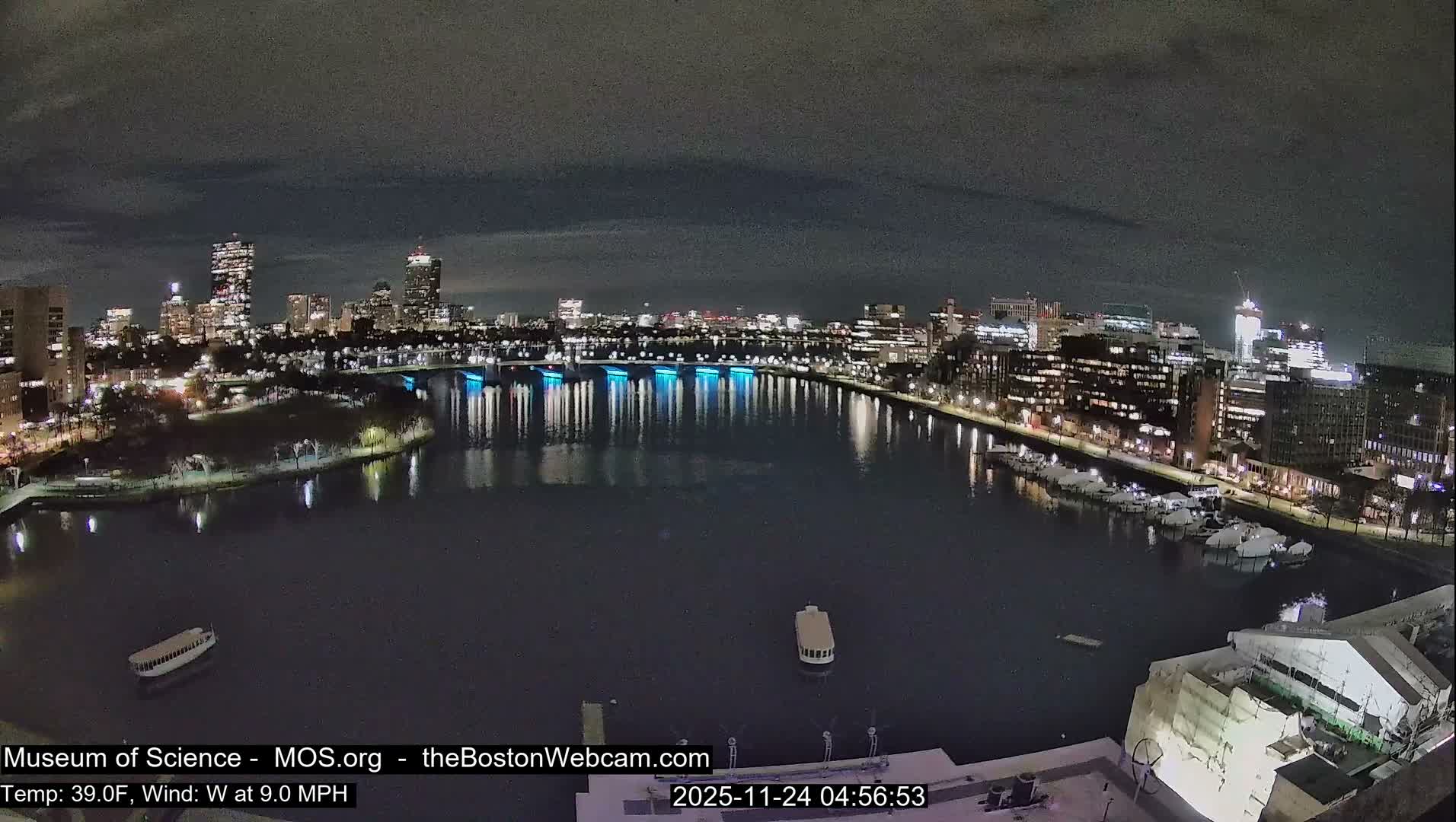 A cloudy night view of a city skyline illuminated across a dark body of water, featuring a blue-lit bridge, numerous city lights reflecting on the water, and several boats, with the temperature at 39°F and a 9 MPH wind.